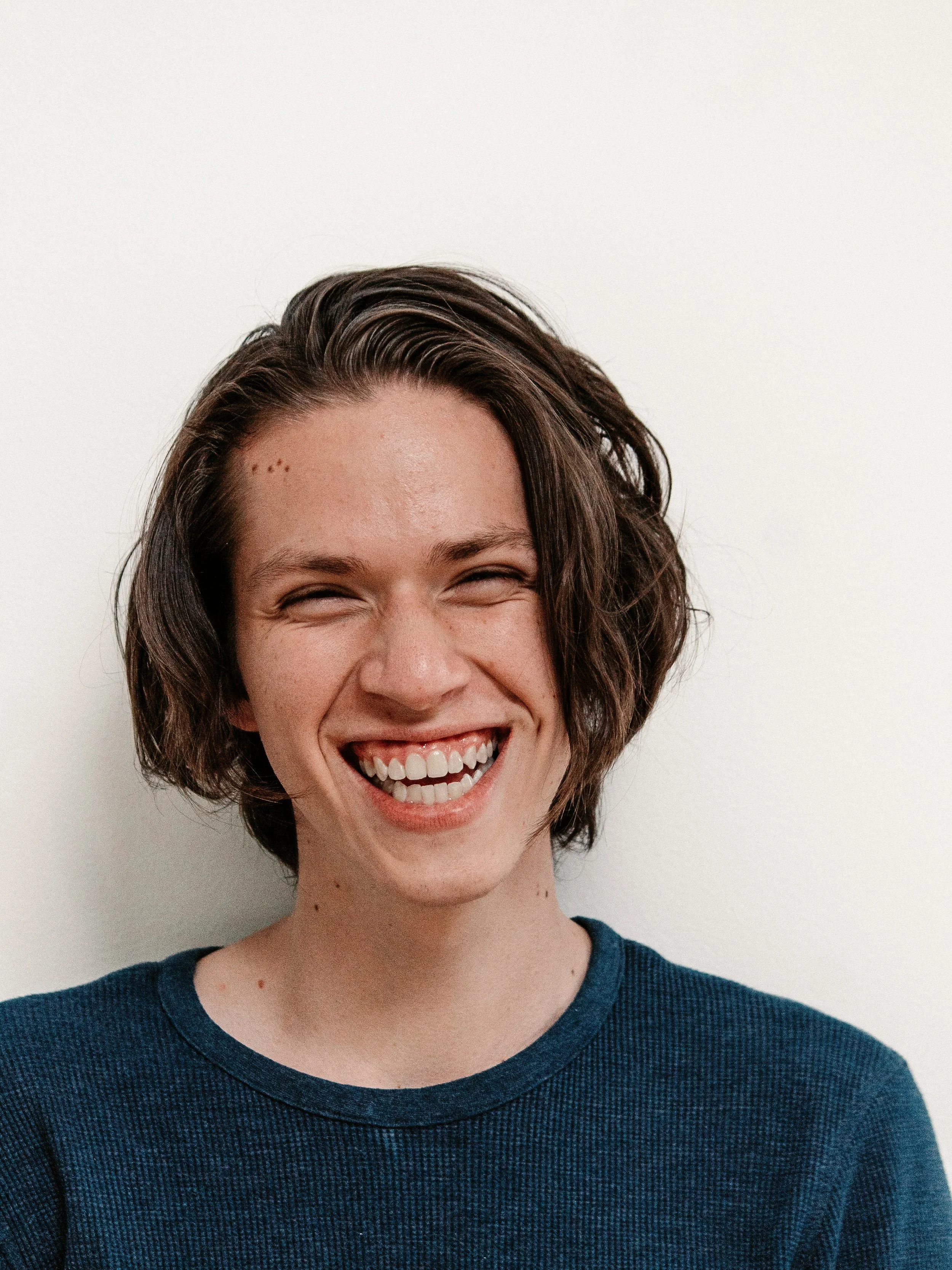A young woman with short wavy brown hair, smiling widely against a plain white background, wearing a dark blue textured shirt.