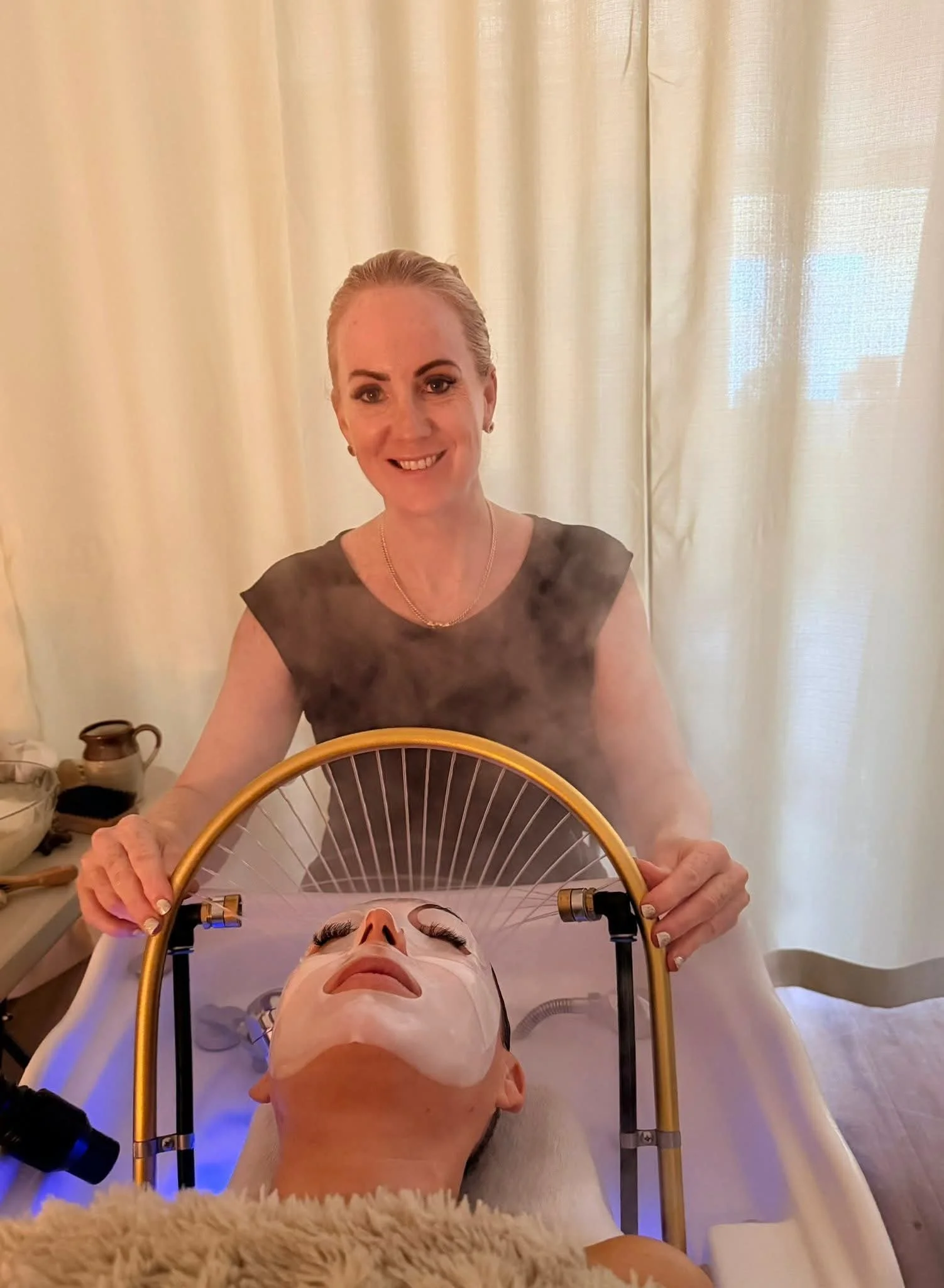 A beauty therapist smiling standing over a person lying down with a facial mask, using a facial steam machine to give a luxurious facial.