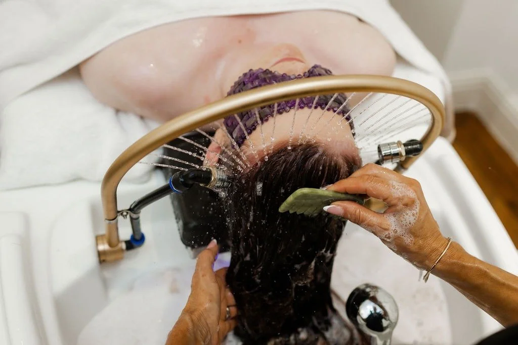 Person receiving a water-based scalp therapy or hair treatment in a salon or spa setting, with a specialized headpiece directing water onto their hair.