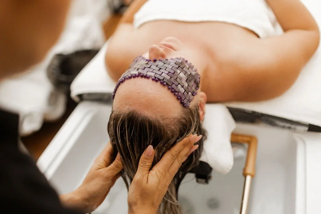 A woman receives a head massage or hair wash at Japanese head spa lying on a salon sink with a purple headrest, while a therapist gently holds her head.