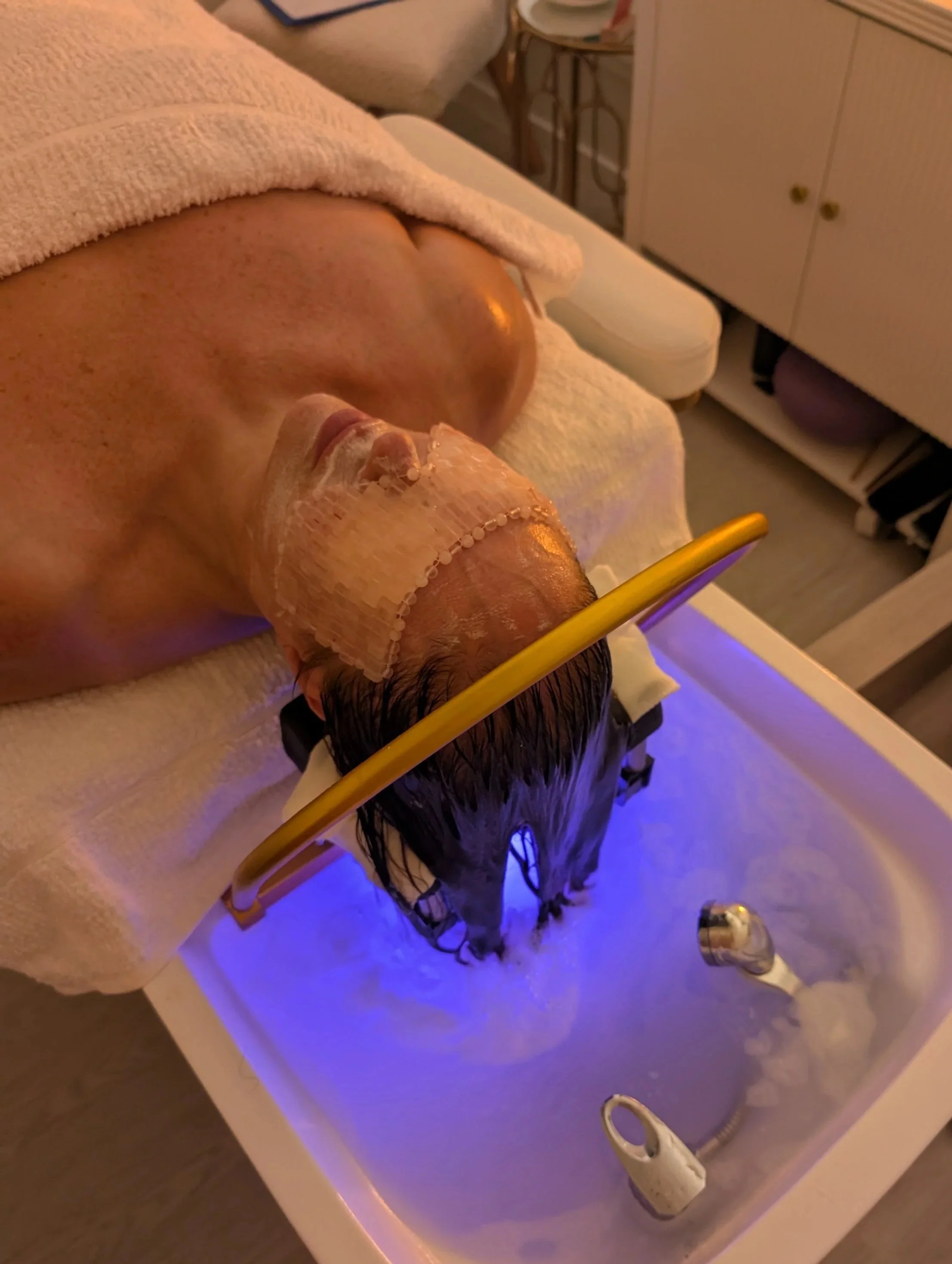 Person receiving a facial treatment while submerged in a blue-lit hydrotherapy tub, with a towel covering their chest and a mask on their face.