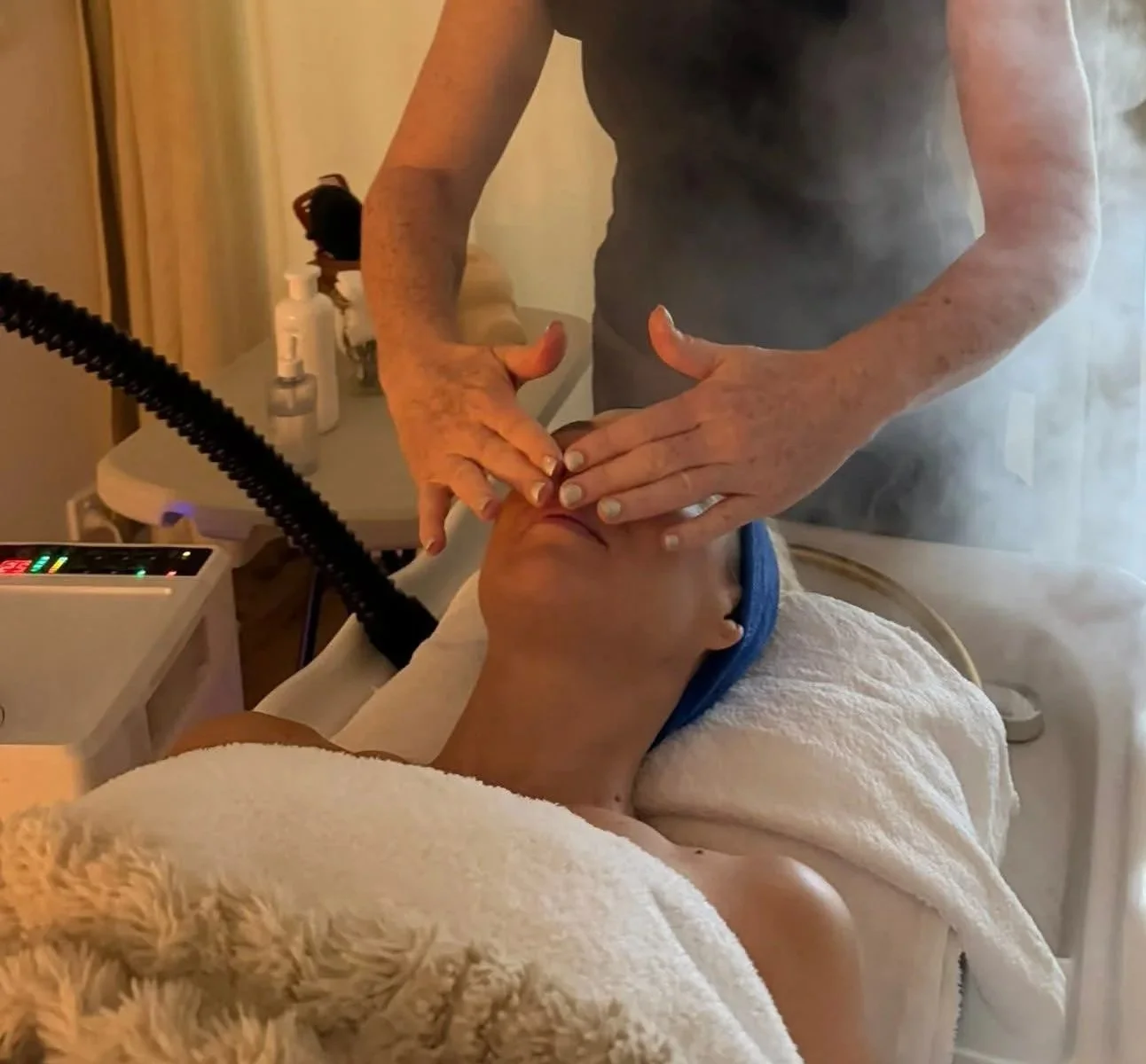 A person receiving a facial treatment from a professional in a spa or clinic setting, with steam visible in the air.
