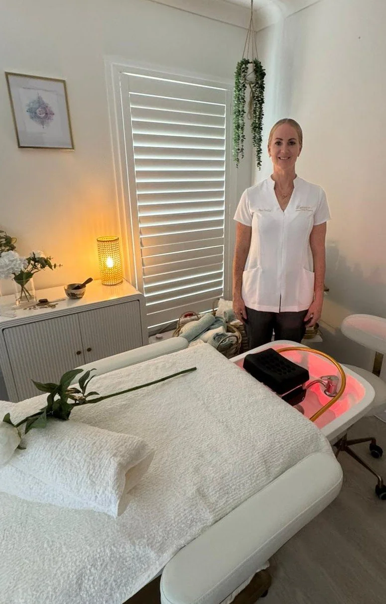 A woman in a white uniform standing in a spa or treatment room with a massage bed, flowers, a candle, and a rolling tray with equipment.