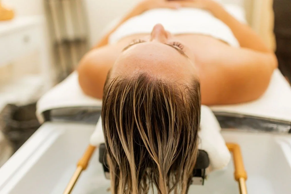 A woman lying on a day spa table during a spa or wellness treatment, with her head tilted back and wet hair hanging down.