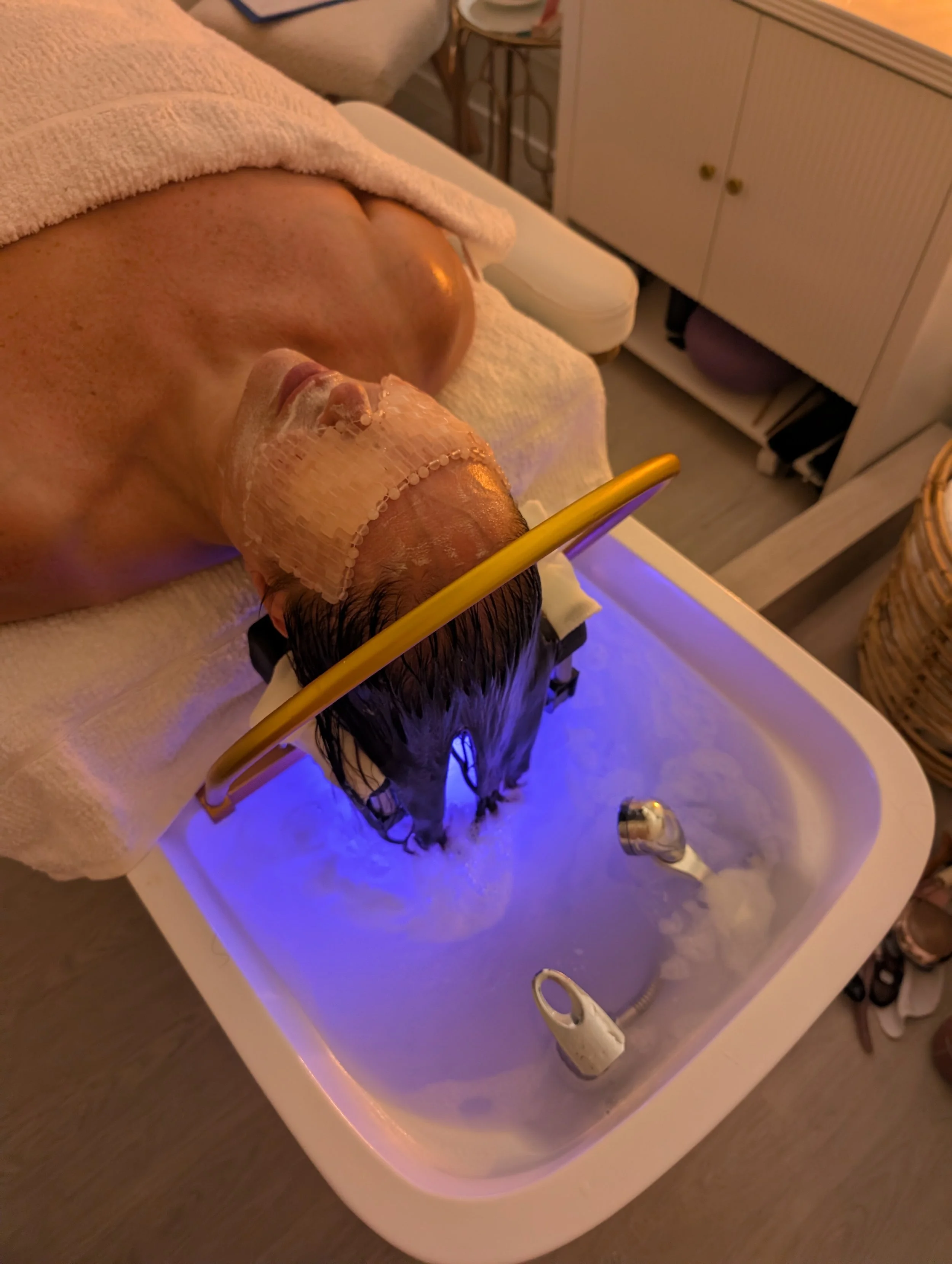 A person receiving a facial treatment or spa therapy in a spa or salon, lying on a treatment bed with a towel over their body, and their face covered with a facial mask or product, with some equipment and furniture visible in the background.