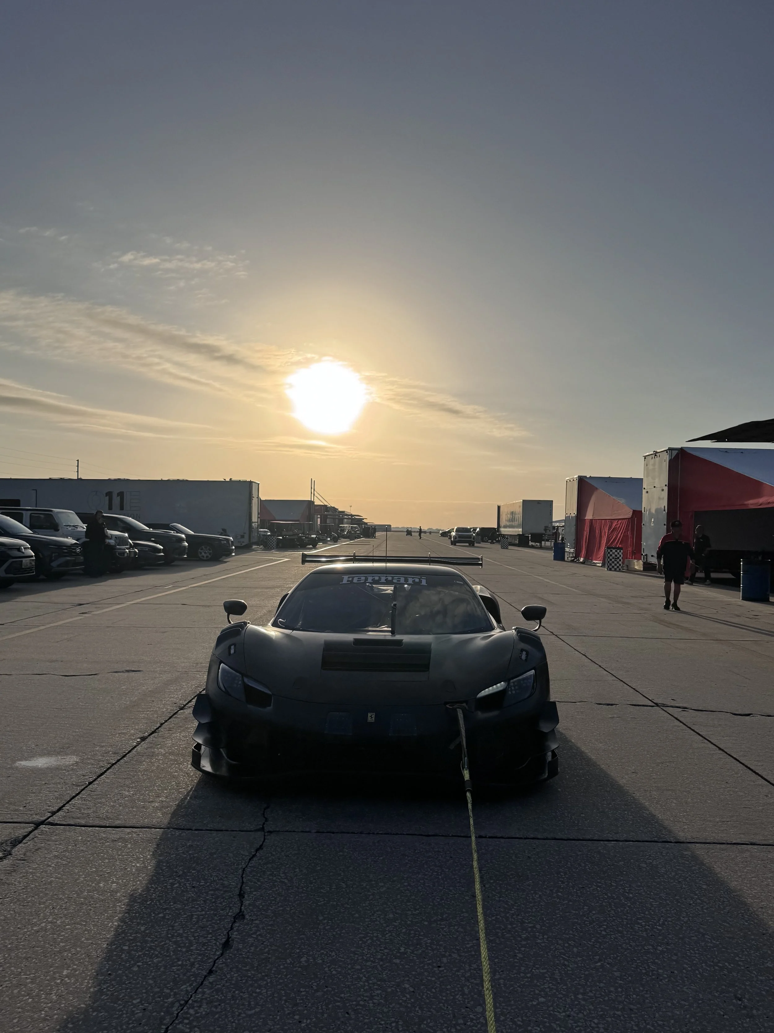 Black Ferrari racing car in a paddock area with trucks and tents, sunset in the background.