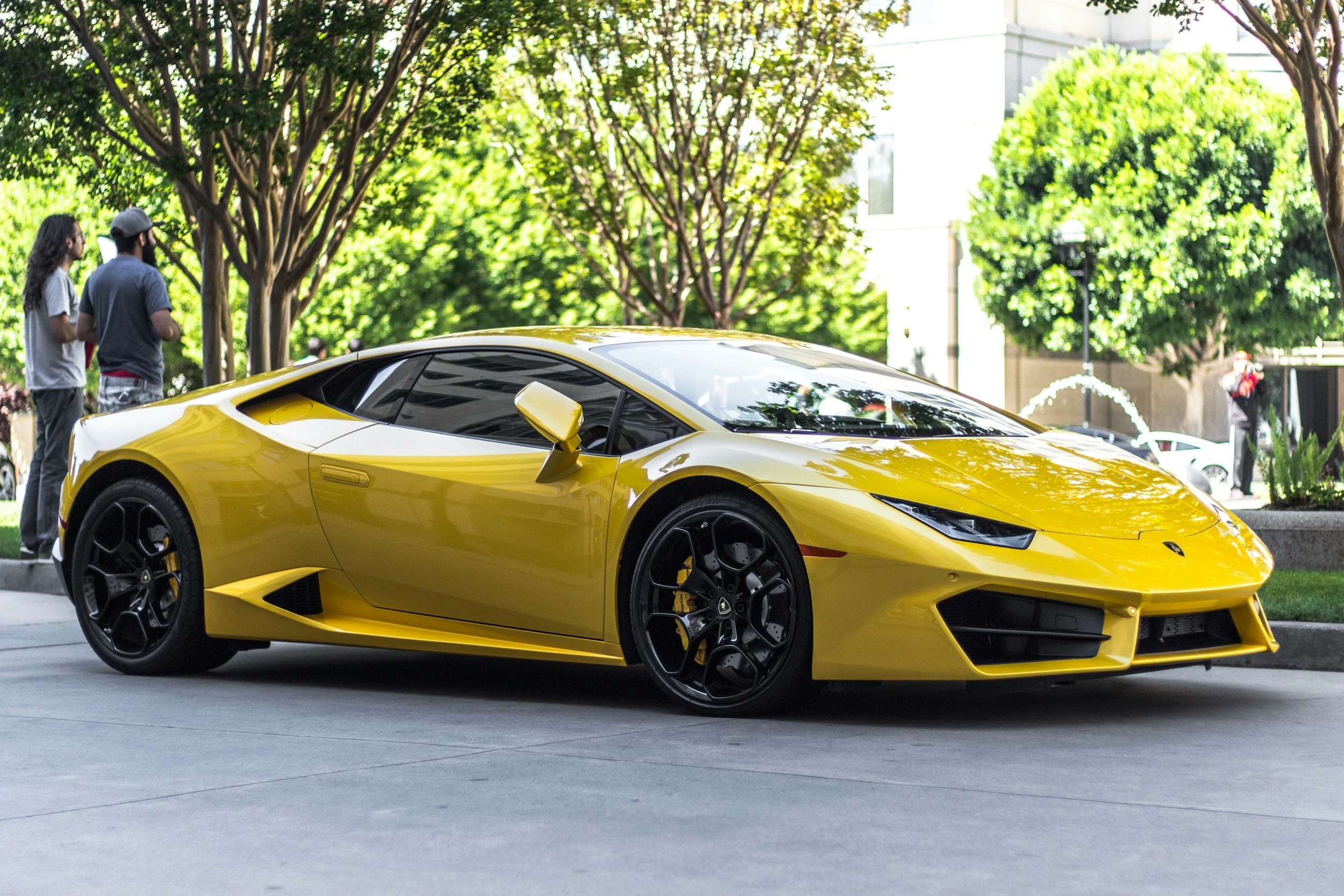 Yellow Lamborghini sports car parked outdoors with people and trees in the background.