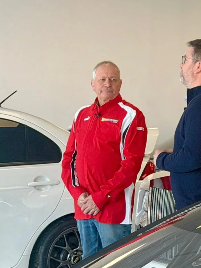 Two men standing near a white car indoors; one man is wearing a red racing jacket and jeans, and the other is in a navy blue sweater with glasses.
