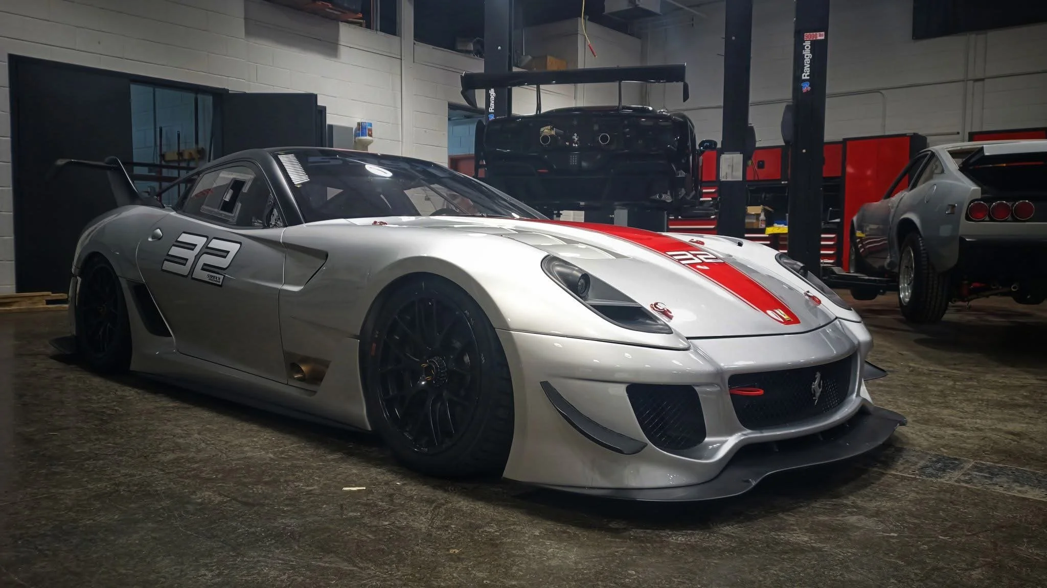 A silver race car with black wheels, racing number 32 on the side, and red and white racing stripes on the hood, parked inside a garage or workshop with other cars and tools in the background.