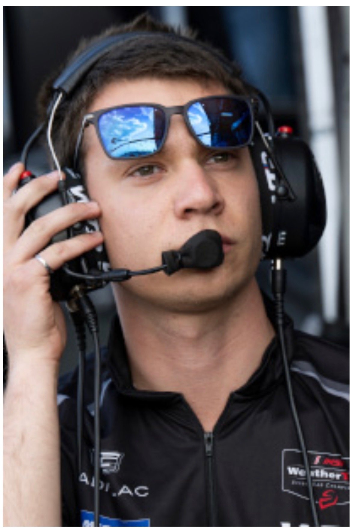 A race car driver wearing sunglasses, a headset with a microphone, and a racing suit, looking serious on the track.