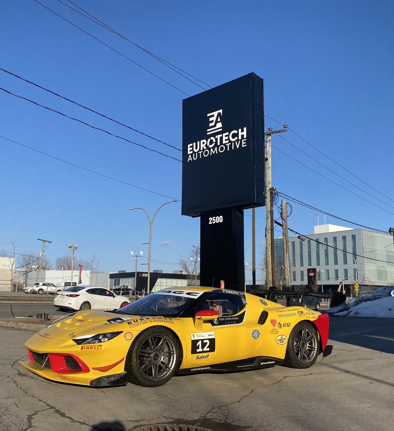 A yellow race car with racing decals parked on a lot in front of a sign for Eurotech Automotive. The car has the number 12 on its door and is surrounded by other vehicles and buildings.