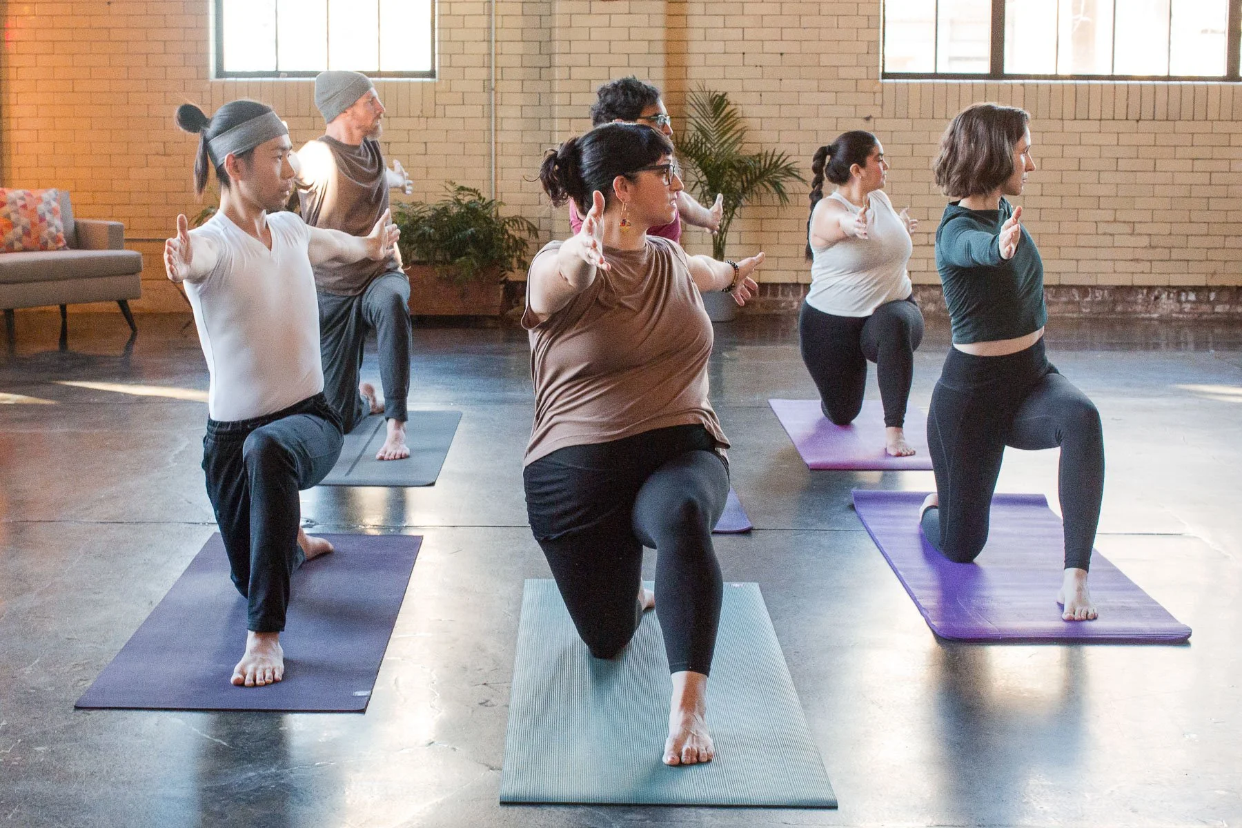 Group of five diverse people participating in a yoga class indoors, kneeling on yoga mats in a spacious, well-lit room with brick walls and large windows.