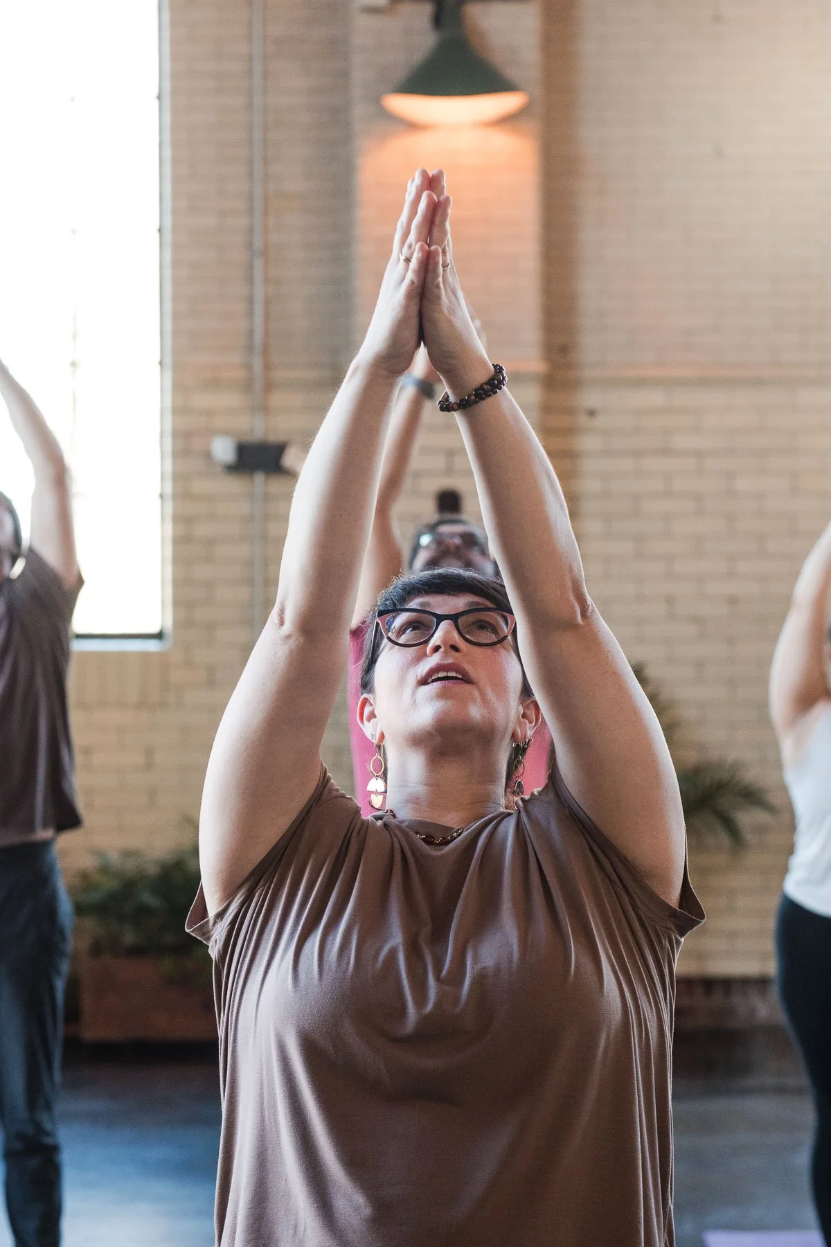 A woman with glasses and earrings is participating in a yoga class, raising her hands in prayer position overhead, with other participants in the background.