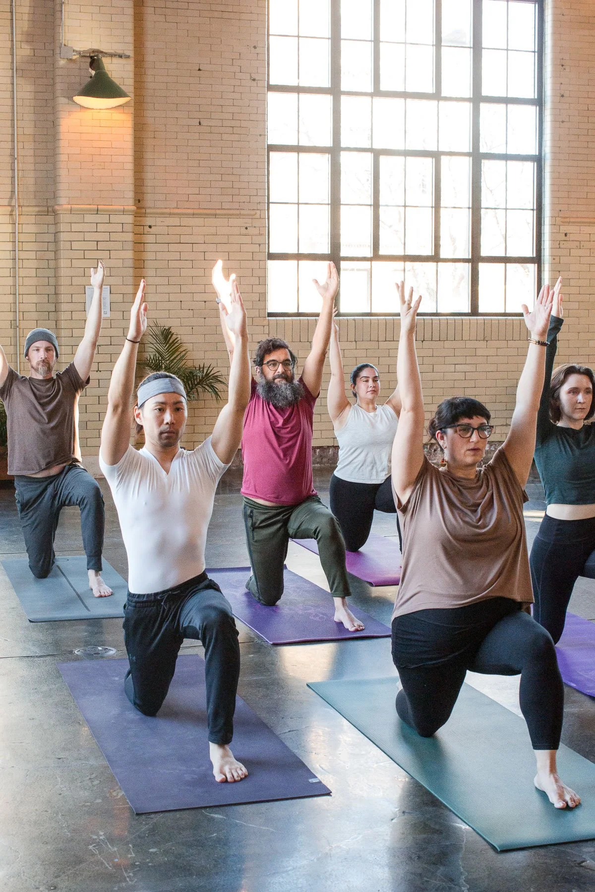 People participating in a yoga class in an industrial-style studio with large windows, raising their arms while kneeling on yoga mats.