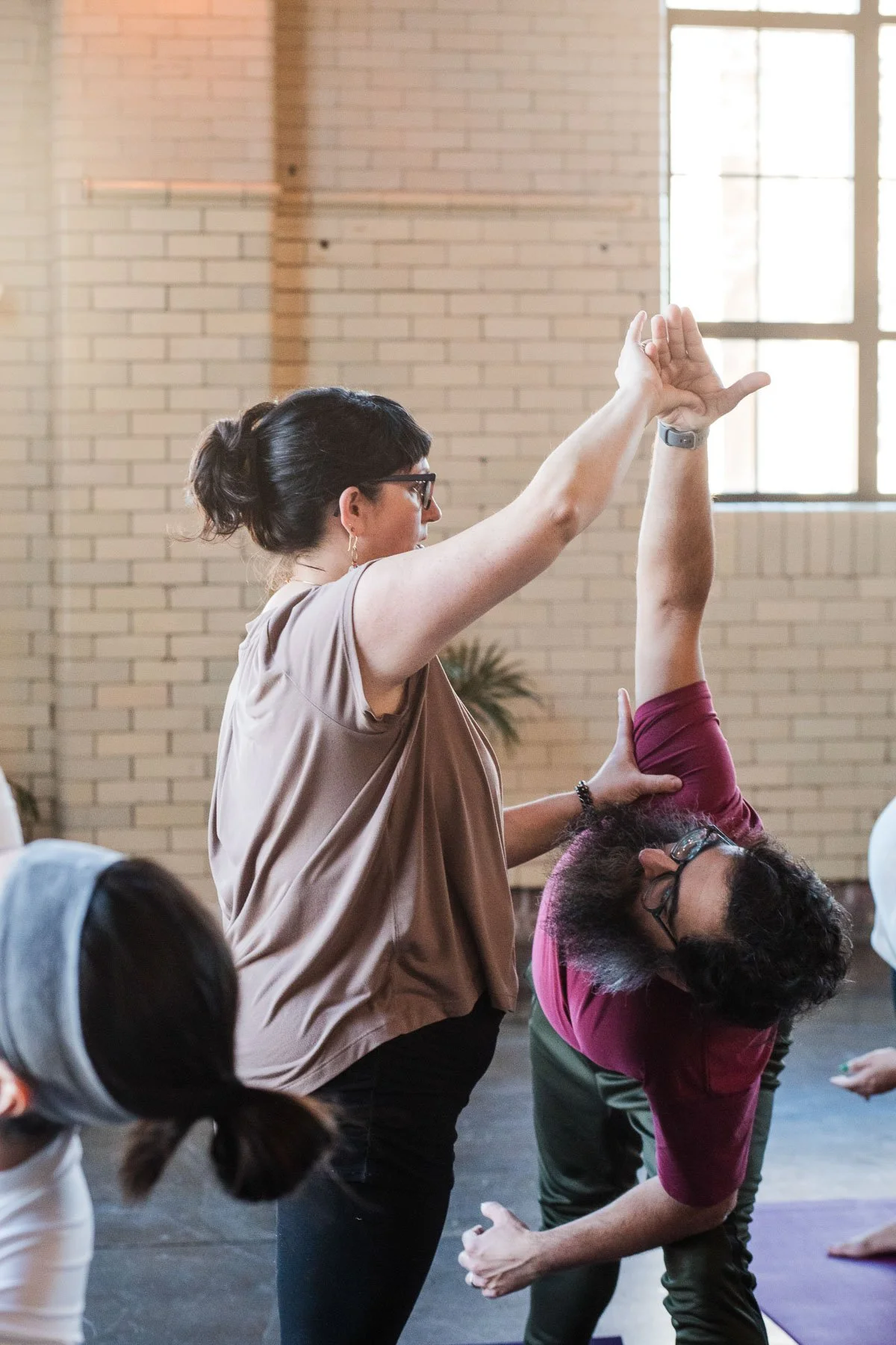 A woman demonstrating a yoga pose with an instructor assisting her in a bright room with large windows and brick walls.