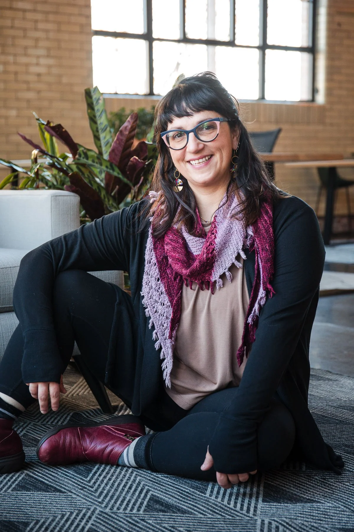 A woman with glasses sitting on the floor, smiling, wearing a black jacket, beige top, purple and magenta scarf, and red boots, in an indoor space with large windows and plants.
