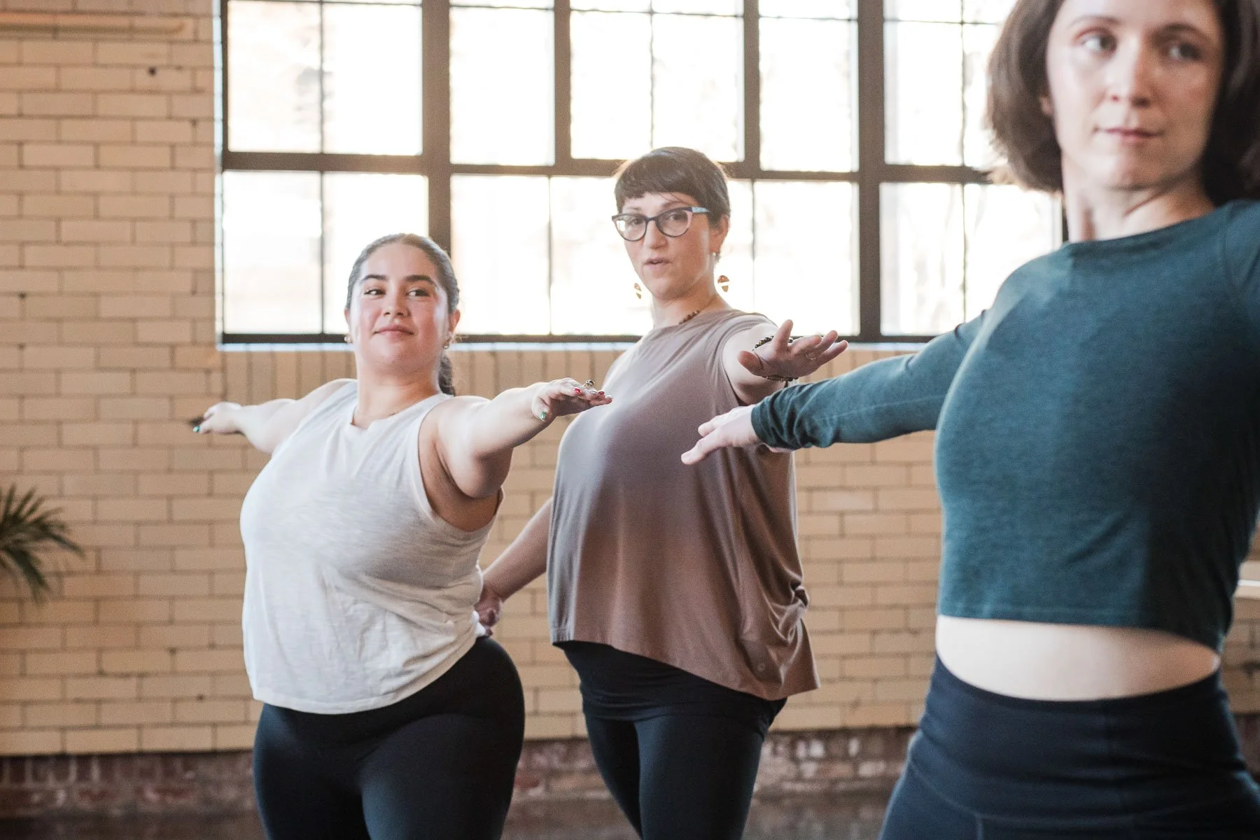 Three women practicing ballet or stretching in a studio with large windows and brick walls.