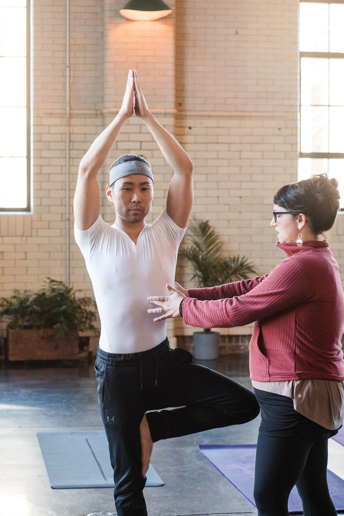 A man practicing yoga in a studio with assistance from a woman, both focused. The man is standing on one leg with palms together above his head, wearing a white shirt and black pants. The woman, wearing glasses and a red jacket, supports him with her