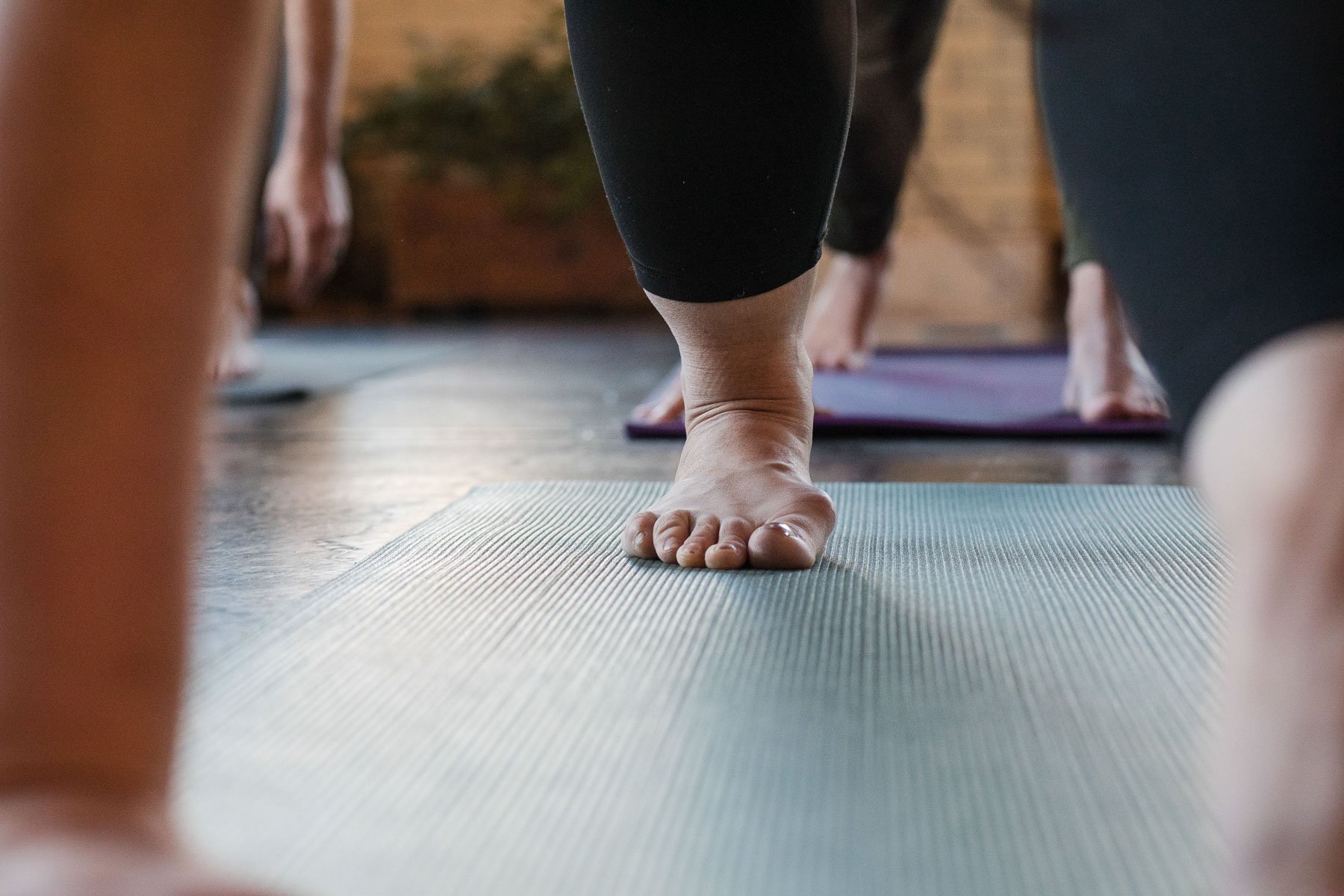 Close-up of a person's foot in a yoga pose on a mat, with other people practicing yoga in the background.
