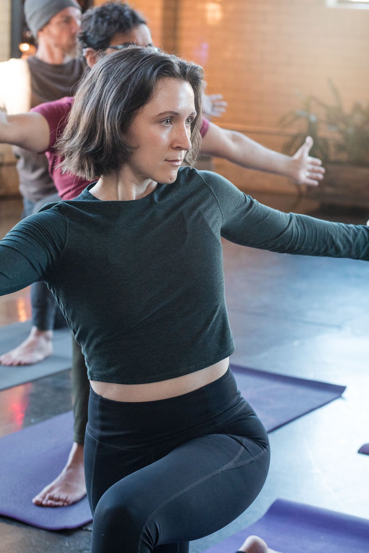 A woman participating in a yoga class, kneeling with arms extended, in a studio with wooden walls and other participants in the background.