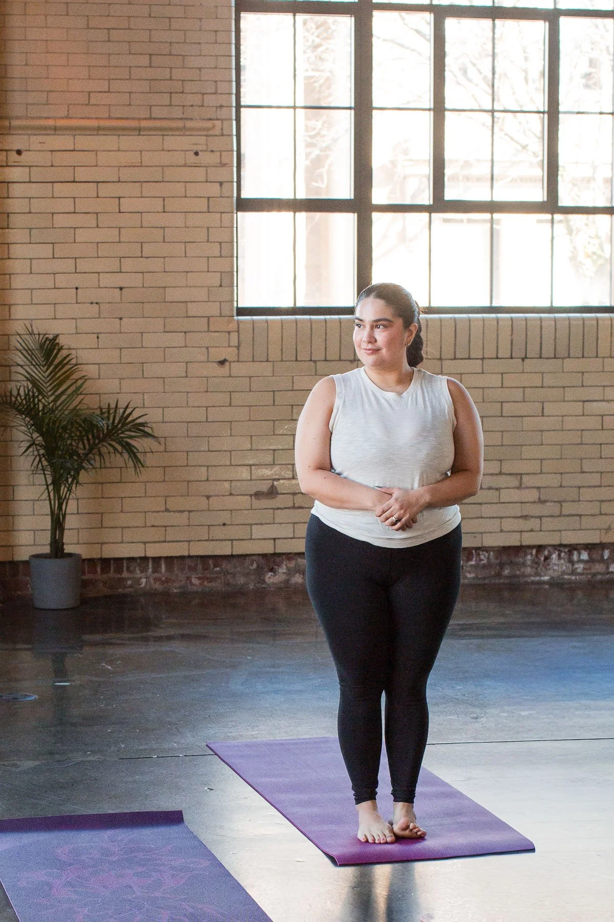 A woman in workout clothes standing on a yoga mat in a spacious room with large windows, brick walls, and a potted plant.