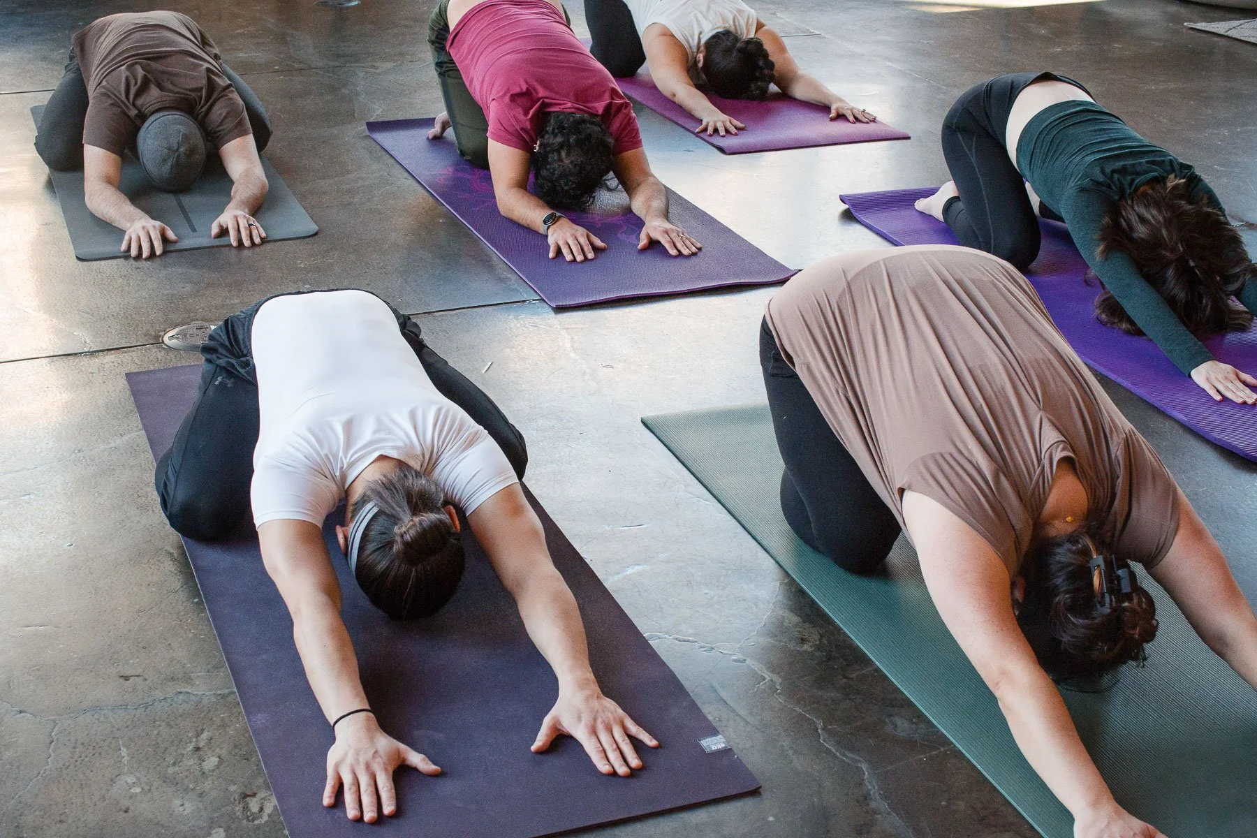 People practicing yoga in a studio, kneeling on mats with their heads bowed and arms extended forward.