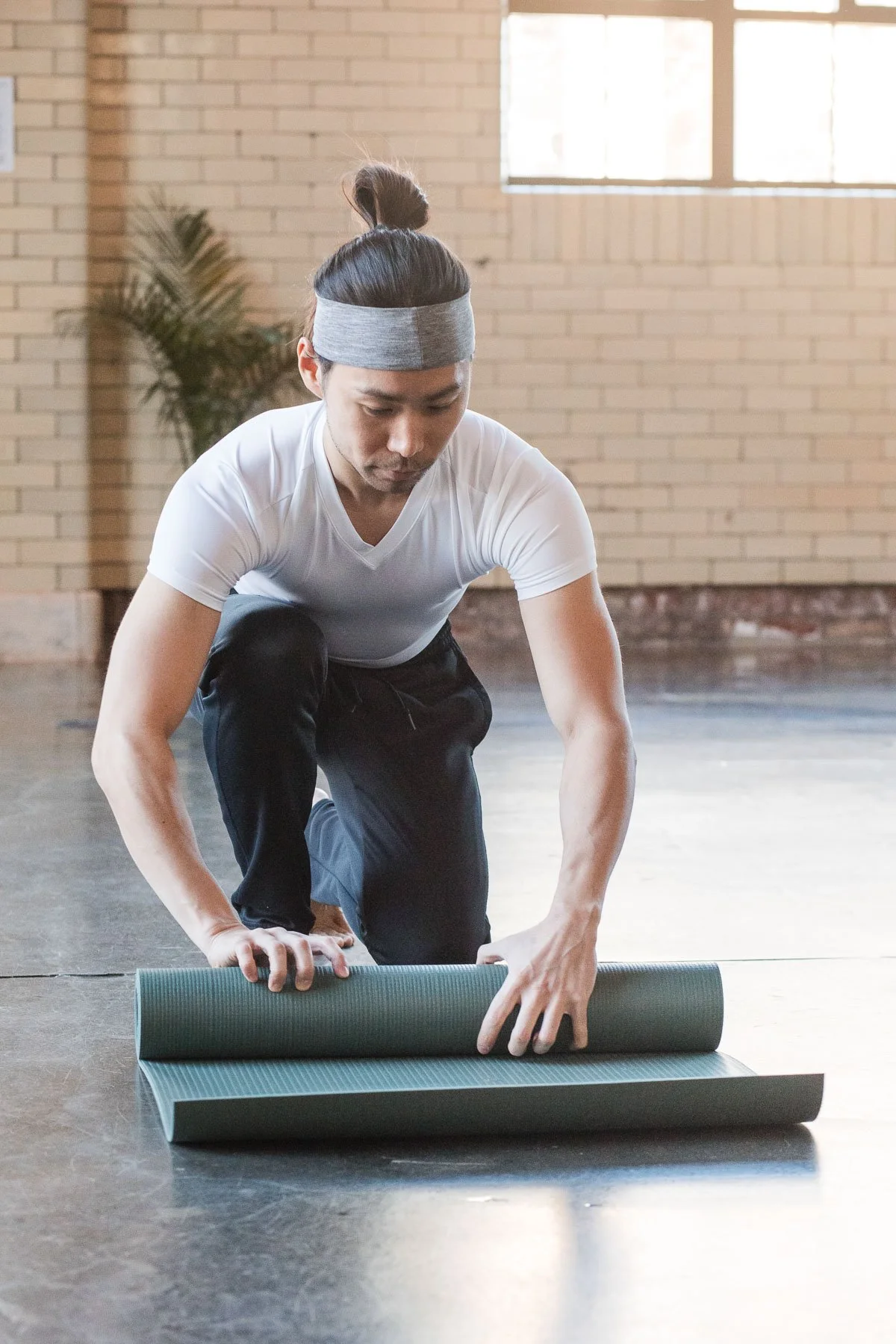 A man in a white t-shirt and black pants kneeling on the floor, rolling out a yoga mat in a brightly lit indoor space with a brick wall and window in the background.