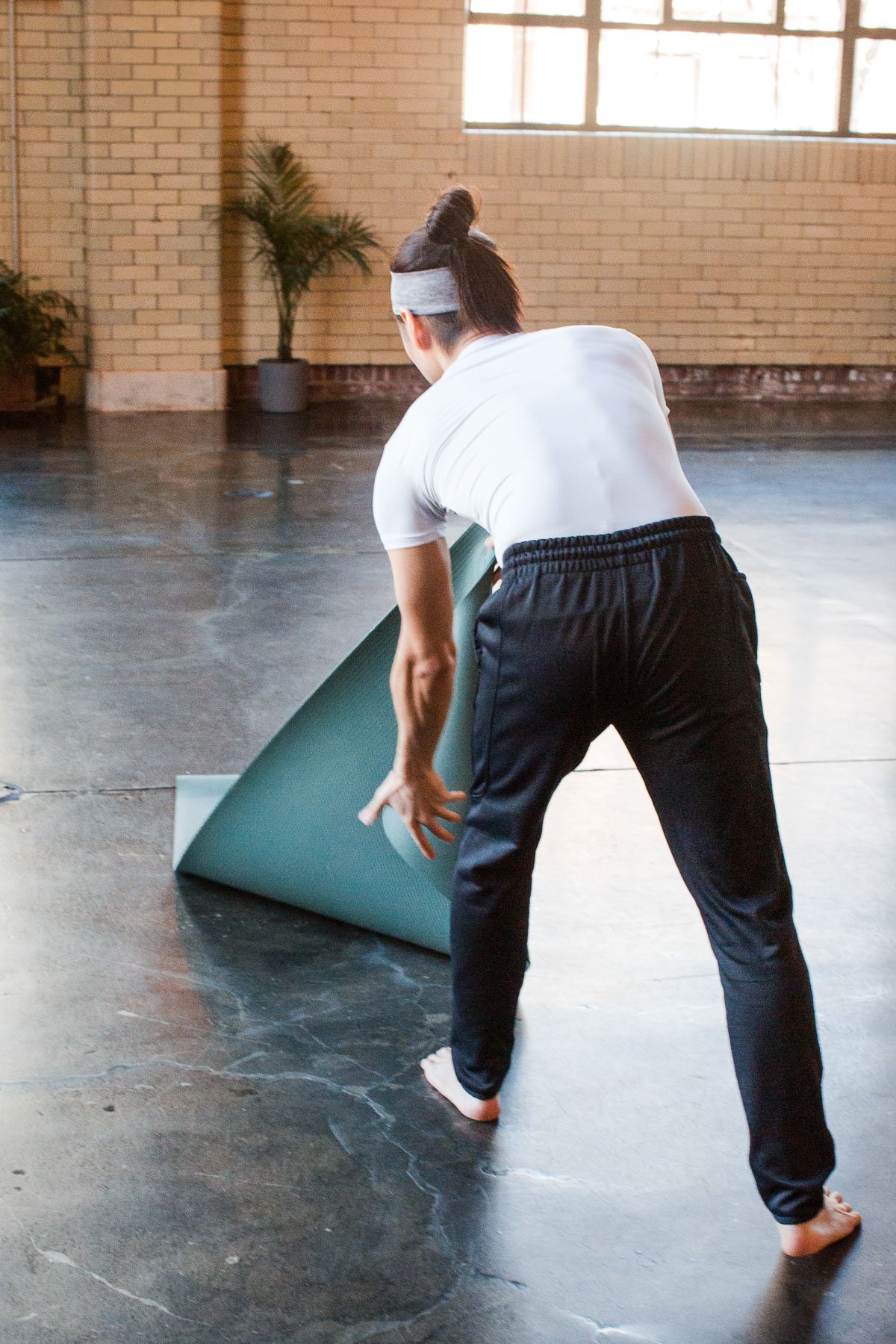 A person, in workout attire, is unfolding a yoga mat on a dark floor in a room with large windows and brick walls.