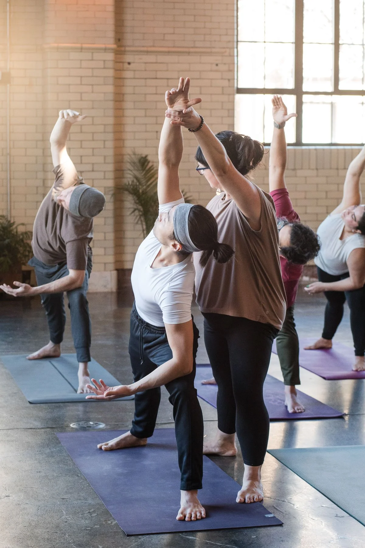 Group of people practicing yoga in a studio, with a teacher assisting a woman in a yoga pose.