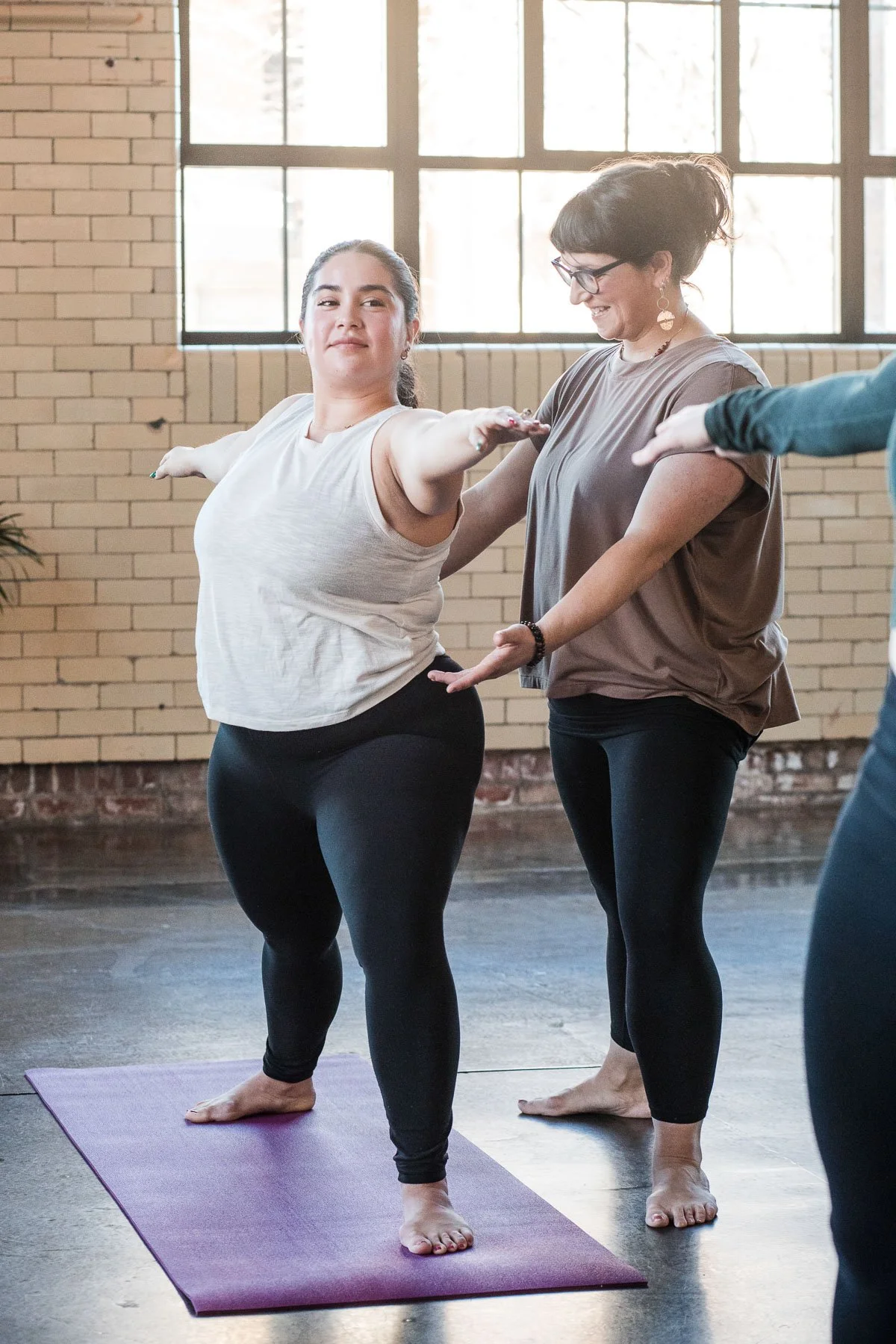 A group of women practicing yoga together indoors, with one woman standing on a yoga mat with her arms outstretched, assisted by another woman.