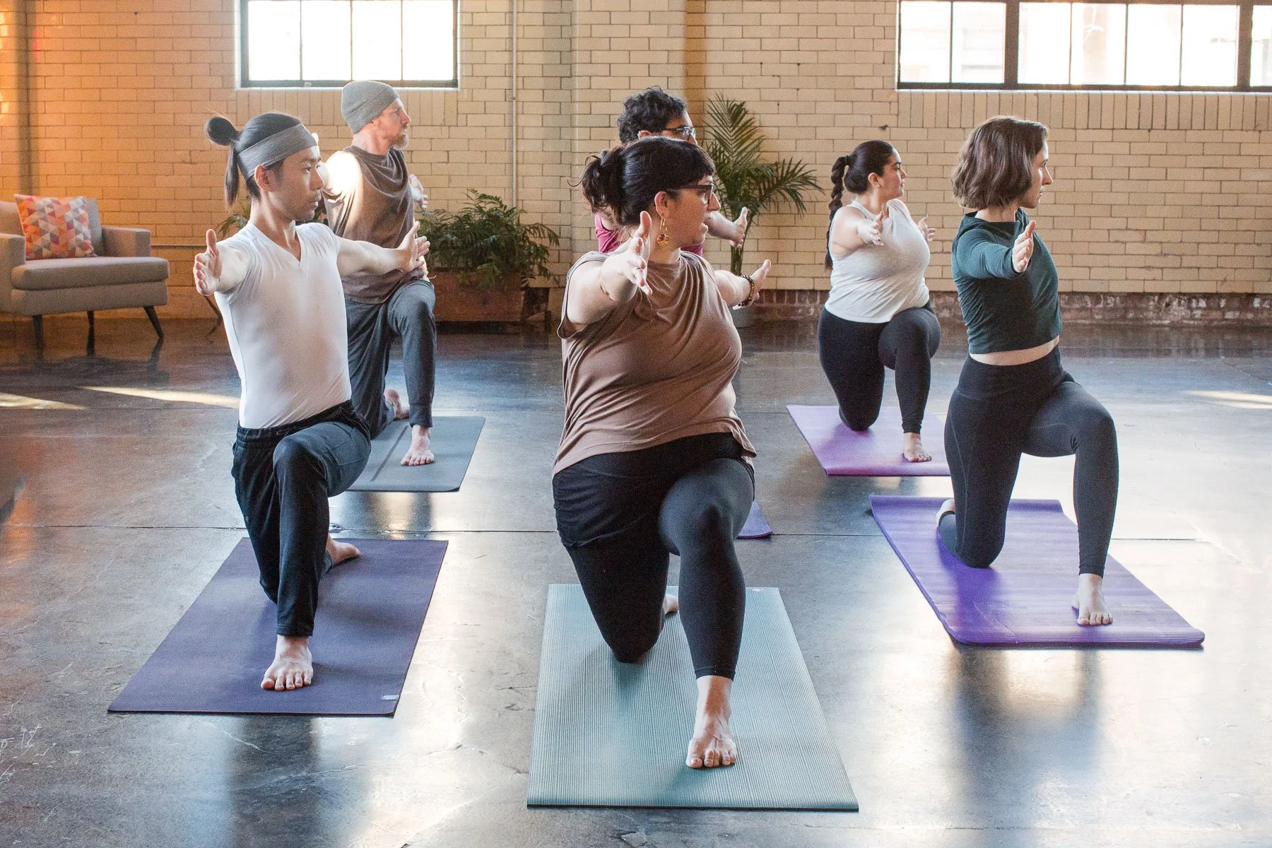 Group of diverse people doing yoga kneeling on mats in a bright indoor studio with large windows and brick walls.