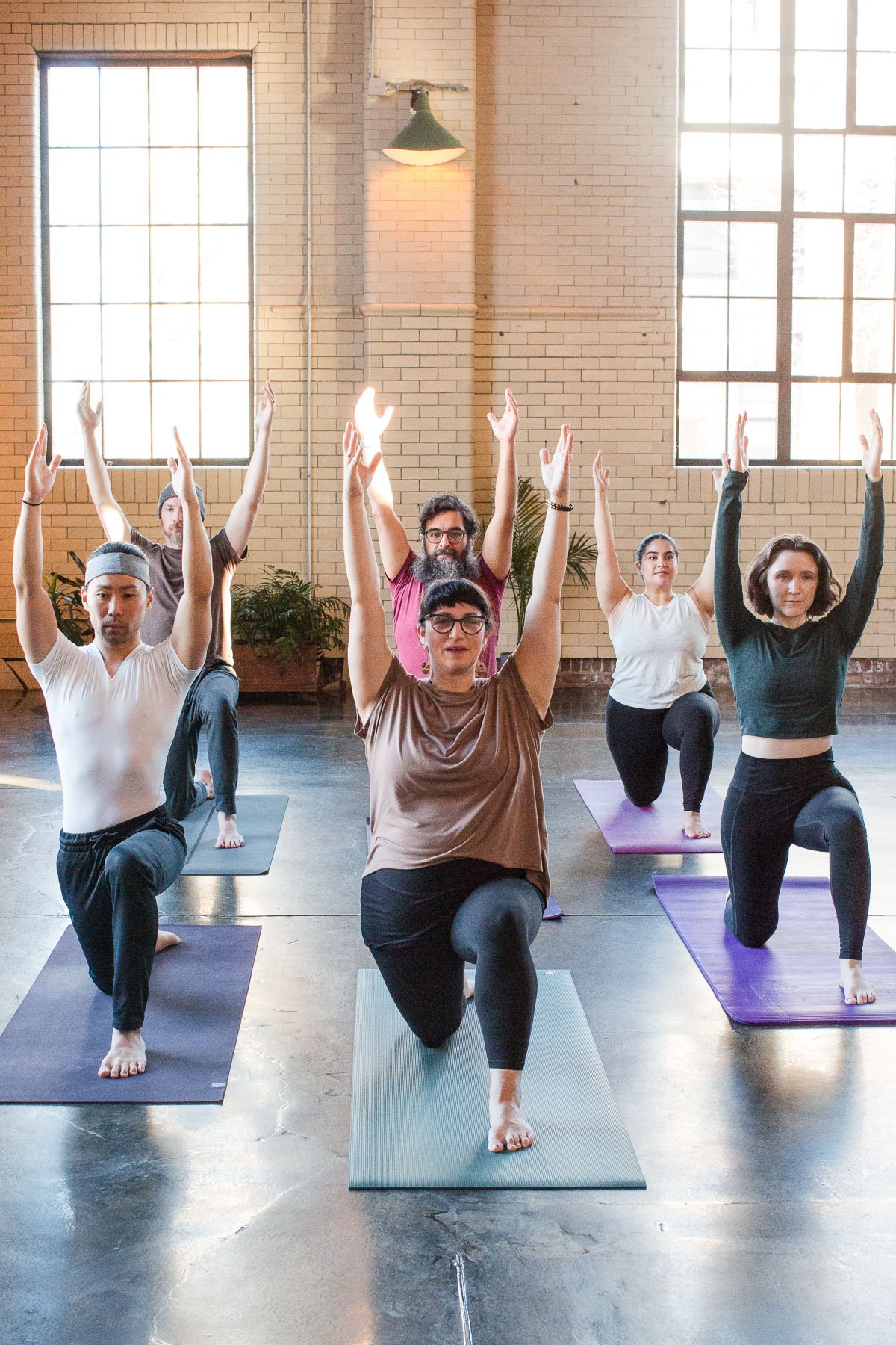 Group of six people practicing yoga in a spacious, well-lit studio with large windows, brick walls, and yoga mats, performing a pose with arms raised.