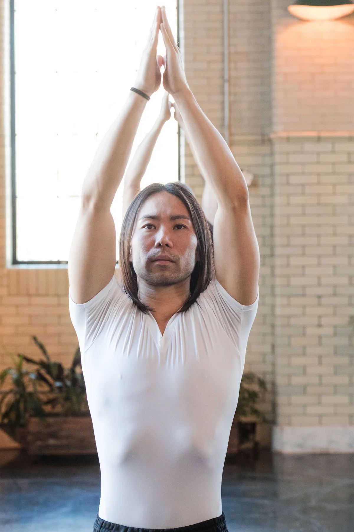 A person with long hair in a white shirt practicing yoga or stretching indoors with arms raised above head and palms pressed together
