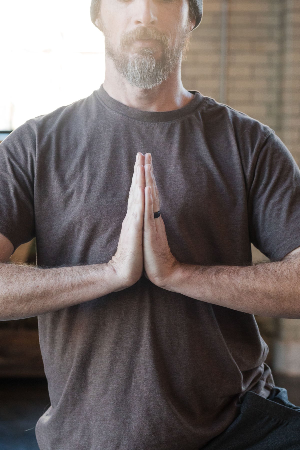A man with a beard wearing a gray t-shirt practicing yoga or meditation, holding his hands together in a prayer position in front of his chest.