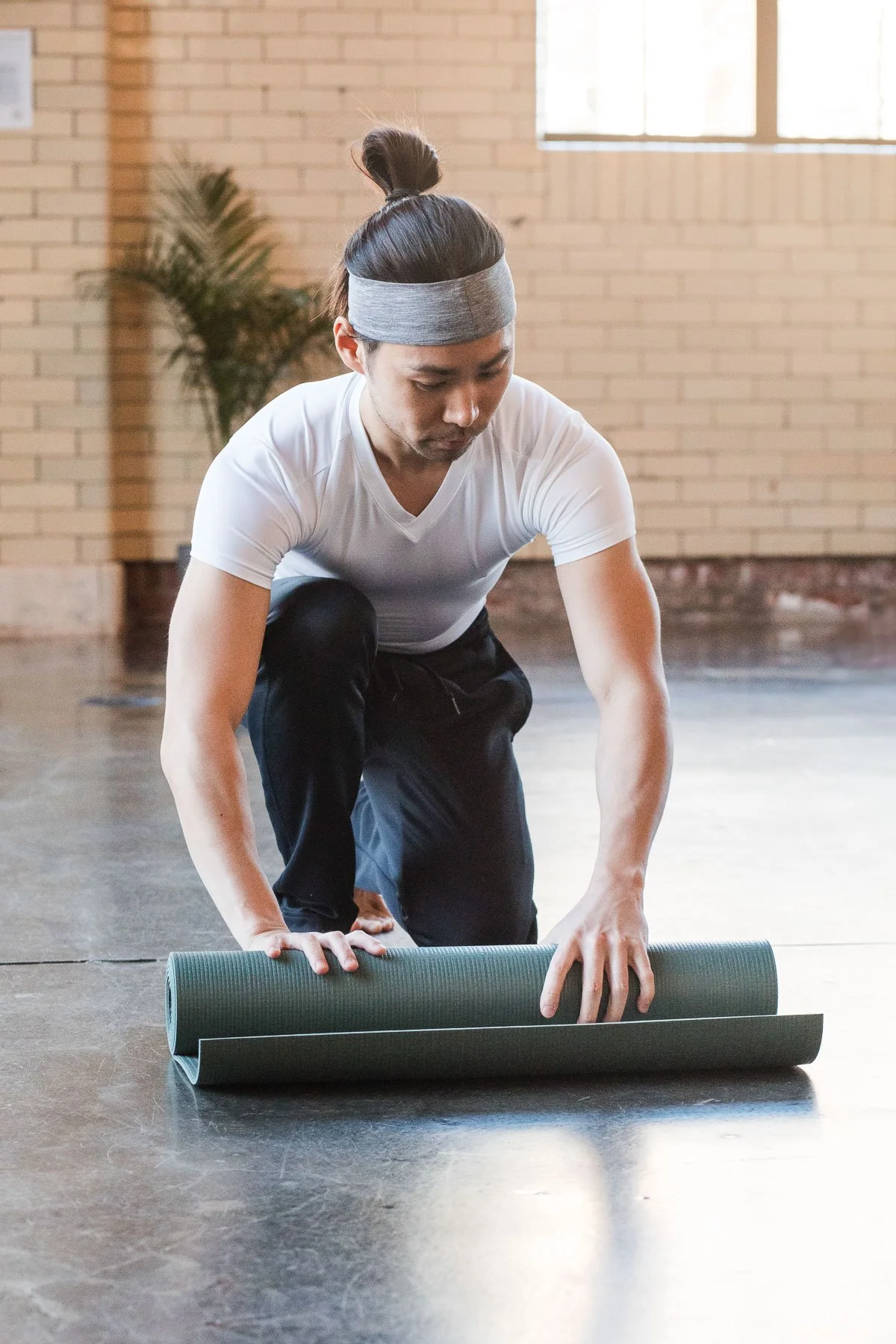 A man with a gray headband and white t-shirt is rolling out a yoga mat on a wooden floor in a gym or studio with brick walls and a window in the background.