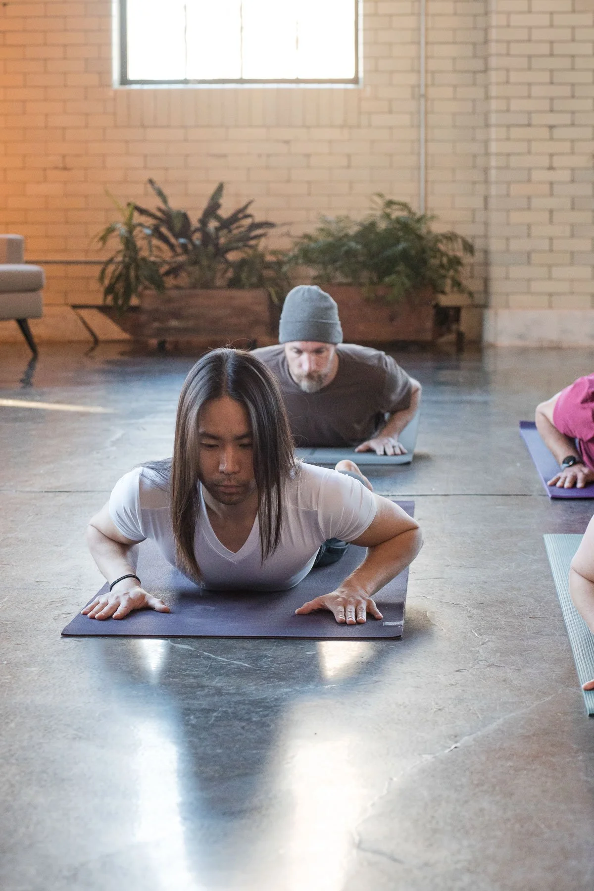 People participating in a yoga class doing forearm plank poses on yoga mats in a bright studio with brick walls and large windows.