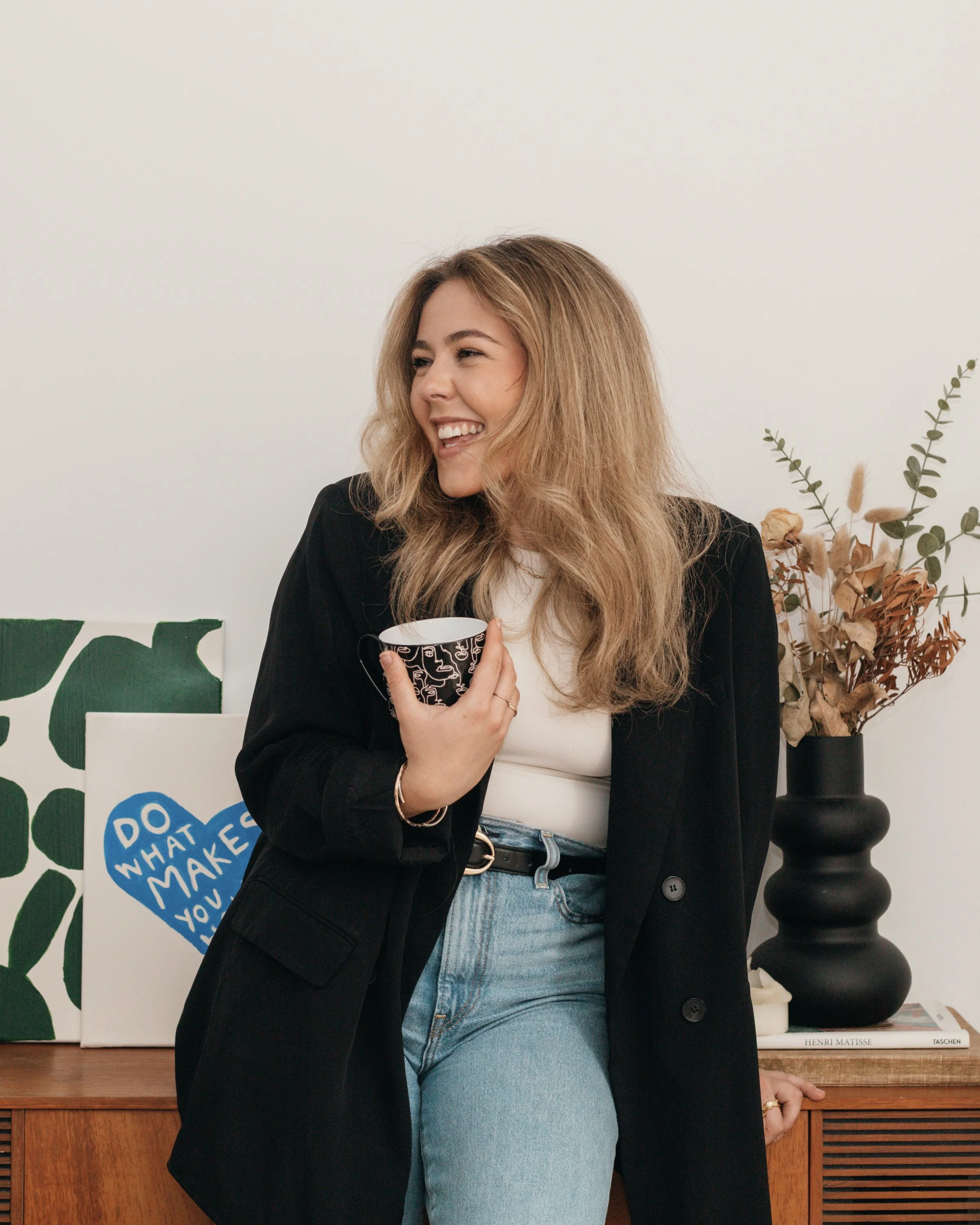 A woman with blonde hair smiling and holding a black and white mug while standing next to a wooden sideboard decorated with artwork and a black vase with dried flowers.