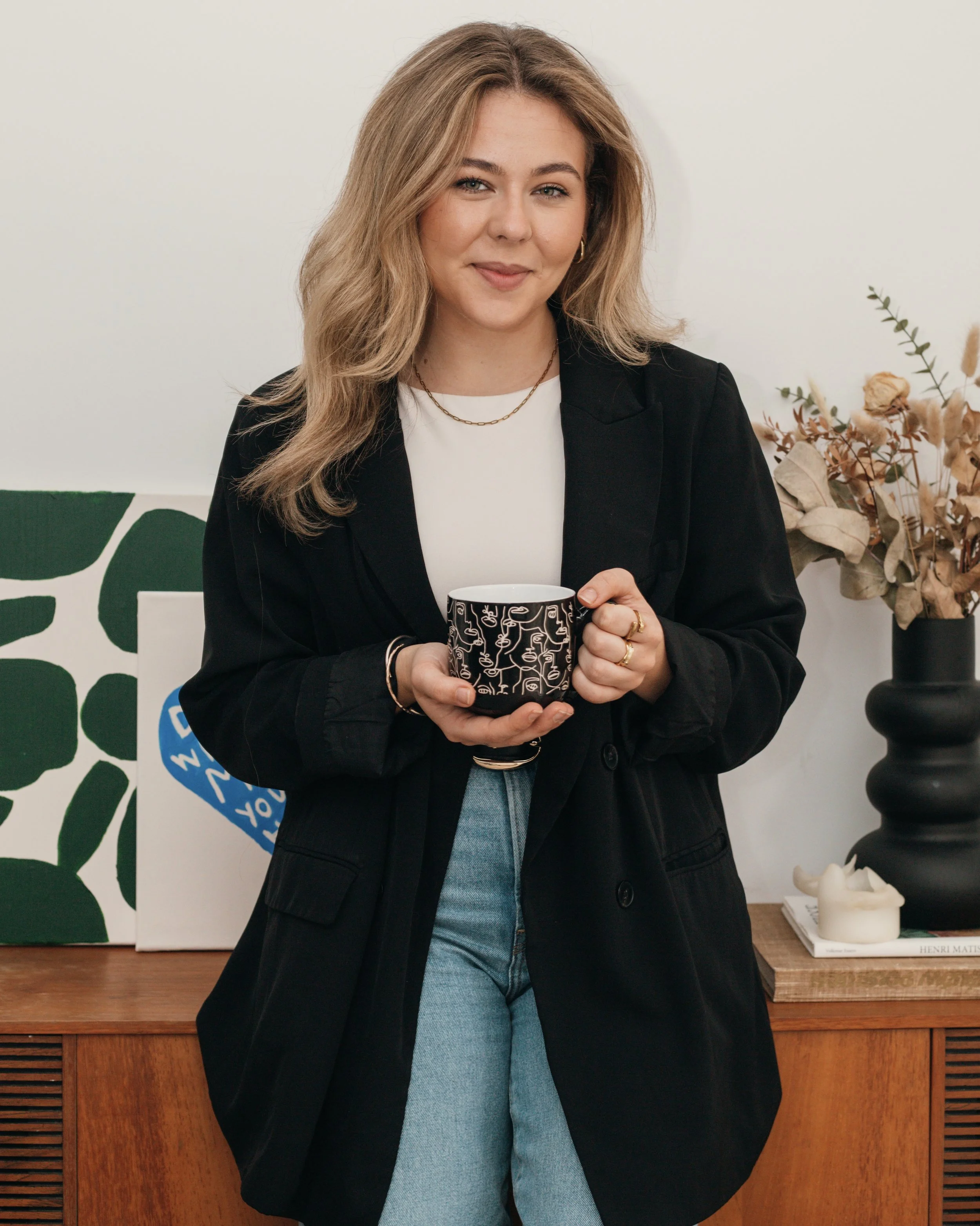 A young woman with shoulder-length blonde hair, wearing a black blazer, white shirt, and light blue jeans, stands indoors holding a black mug with white line drawings, smiling softly, with plants and abstract art in the background.