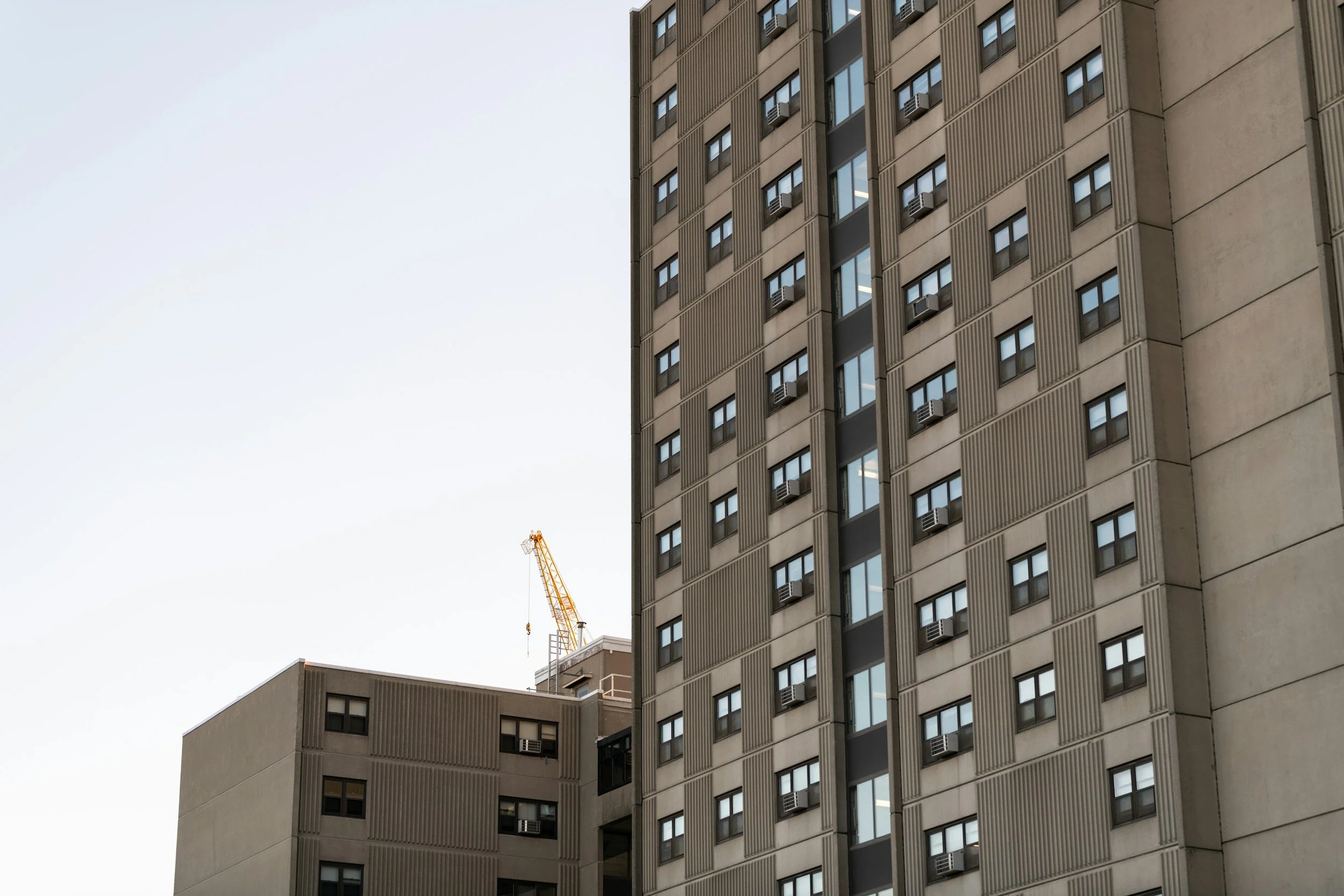 Two high-rise buildings with windows and air conditioning units, with a construction crane in the background under a clear sky.