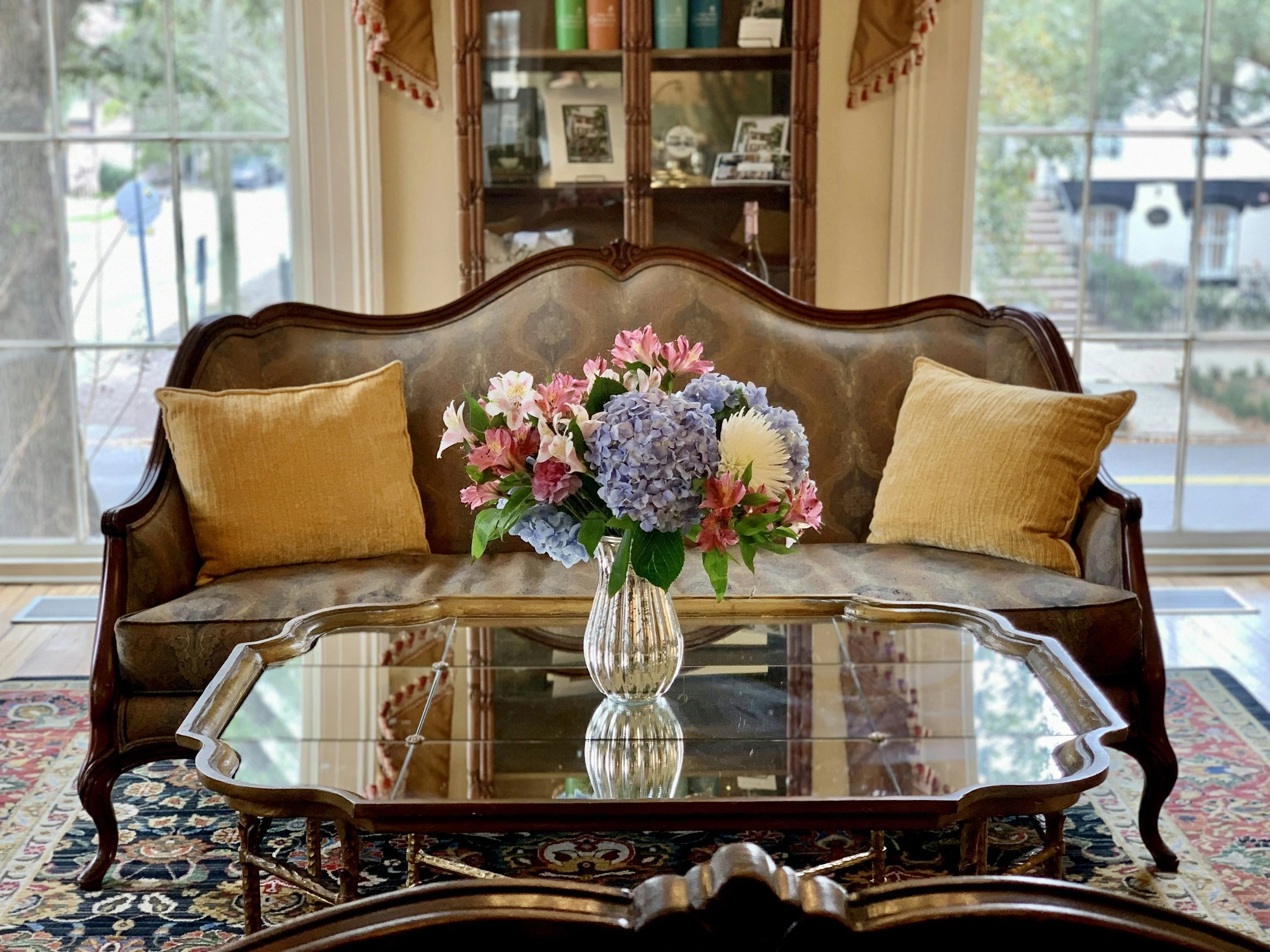 A vintage living room with a wooden sofa, mustard cushions, a glass-topped coffee table, and a vase of pink, purple, and white flowers. The room has large windows with a view of a street and outside houses.