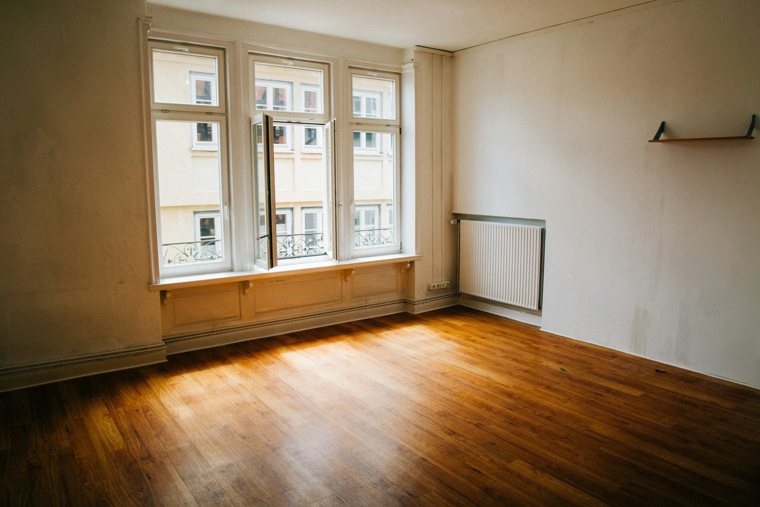 Empty room with hardwood floor, large window with open window panes, radiator, and a small empty wall-mounted shelf.