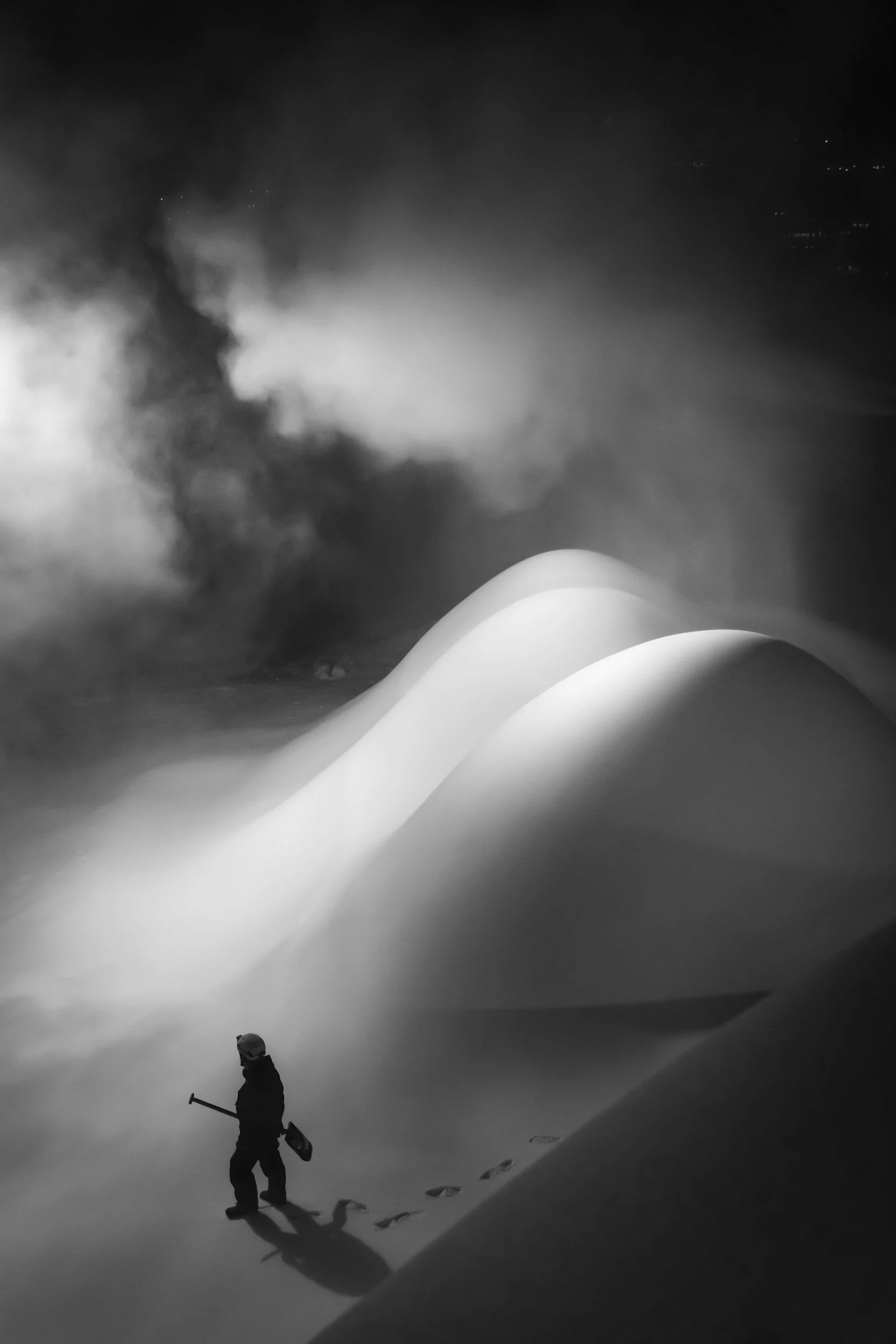A lone snow shoveler walks through a snowy landscape at night, with large snowdrifts and mist or smoke rising in the background, all in black and white.