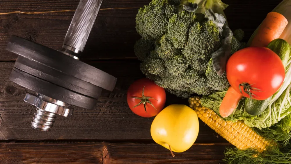 Dumbbell and fresh vegetables on a wooden surface, including tomatoes, broccoli, carrots, yellow squash, corn, and leafy greens.