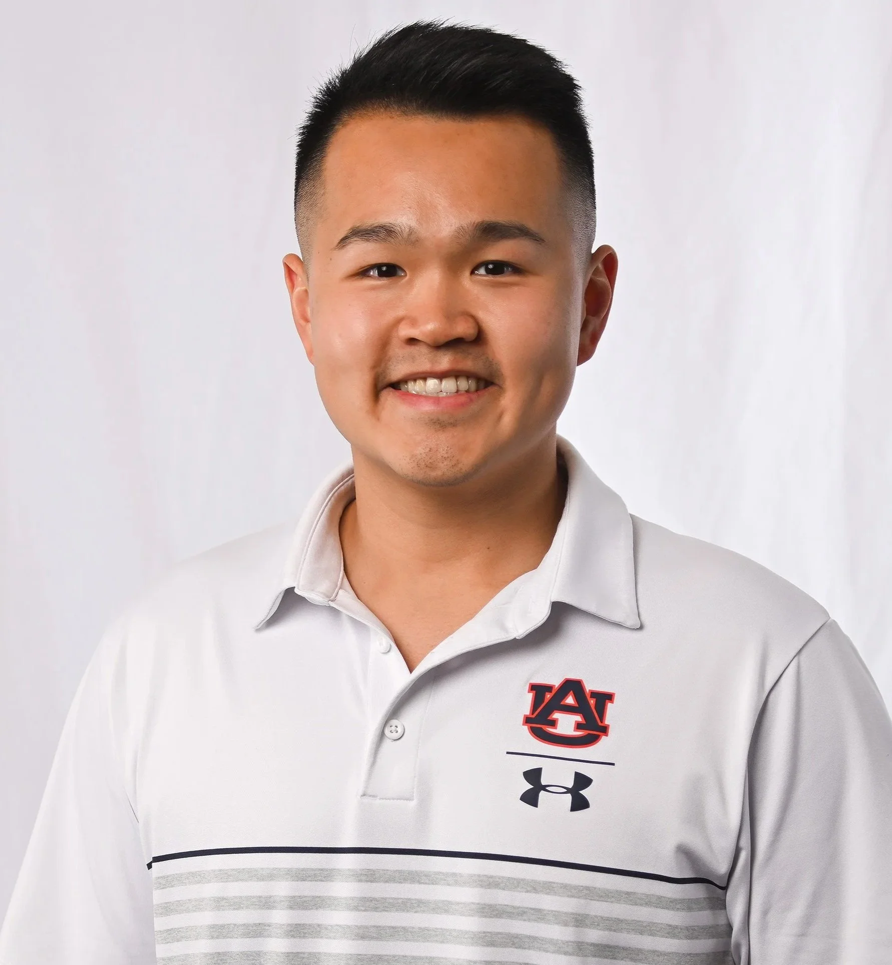 A young Asian man smiling wearing a white sports polo shirt with an Auburn University logo and Under Armour logo, standing against a plain white background.