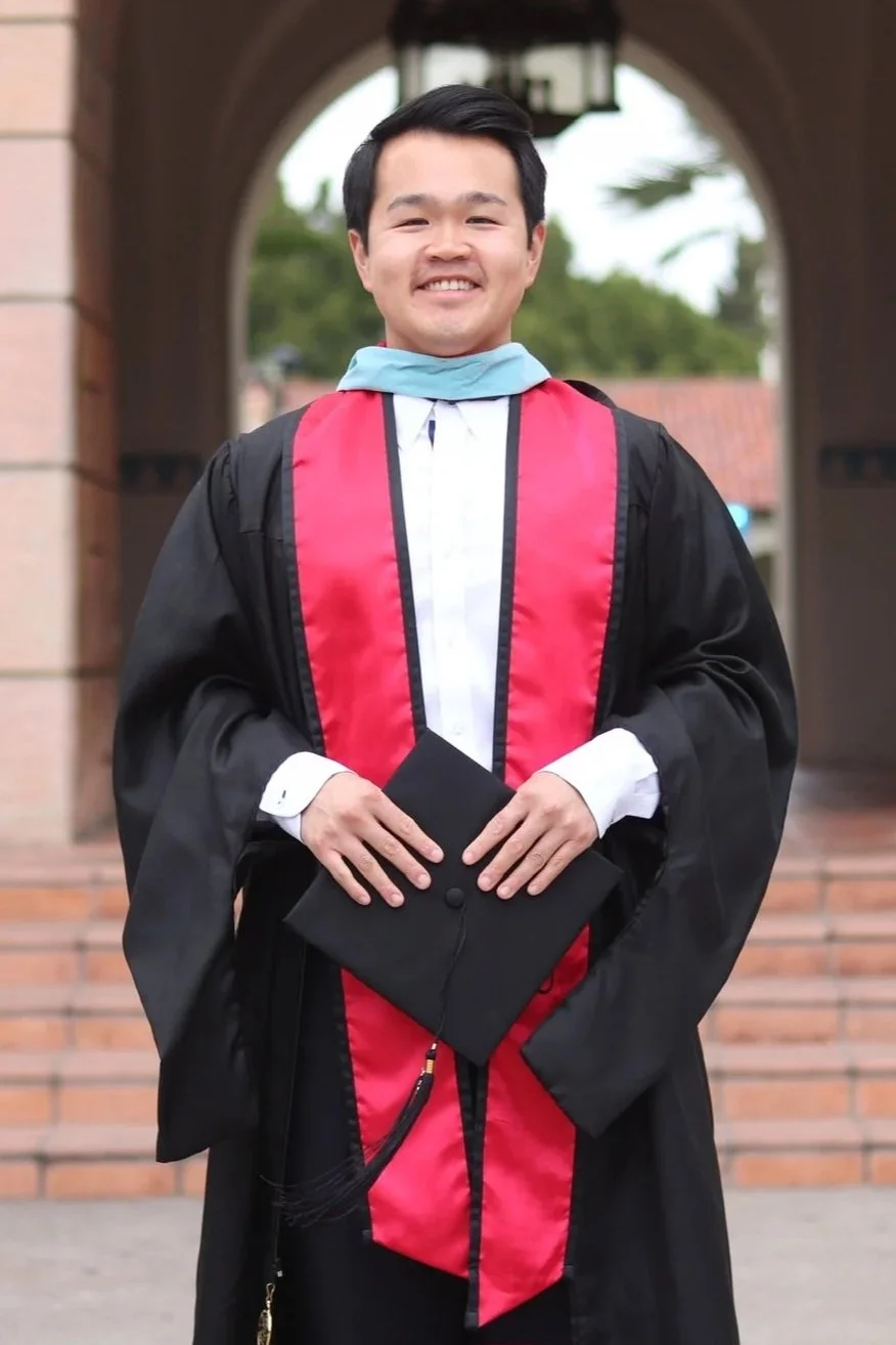 A young man in graduation gown and cap holding a diploma outside near a brick archway.
