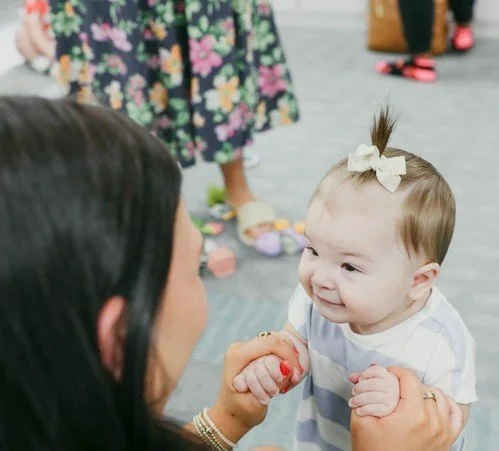 A baby girl with a white bow in her hair smiling and holding hands with an adult woman in a public place.