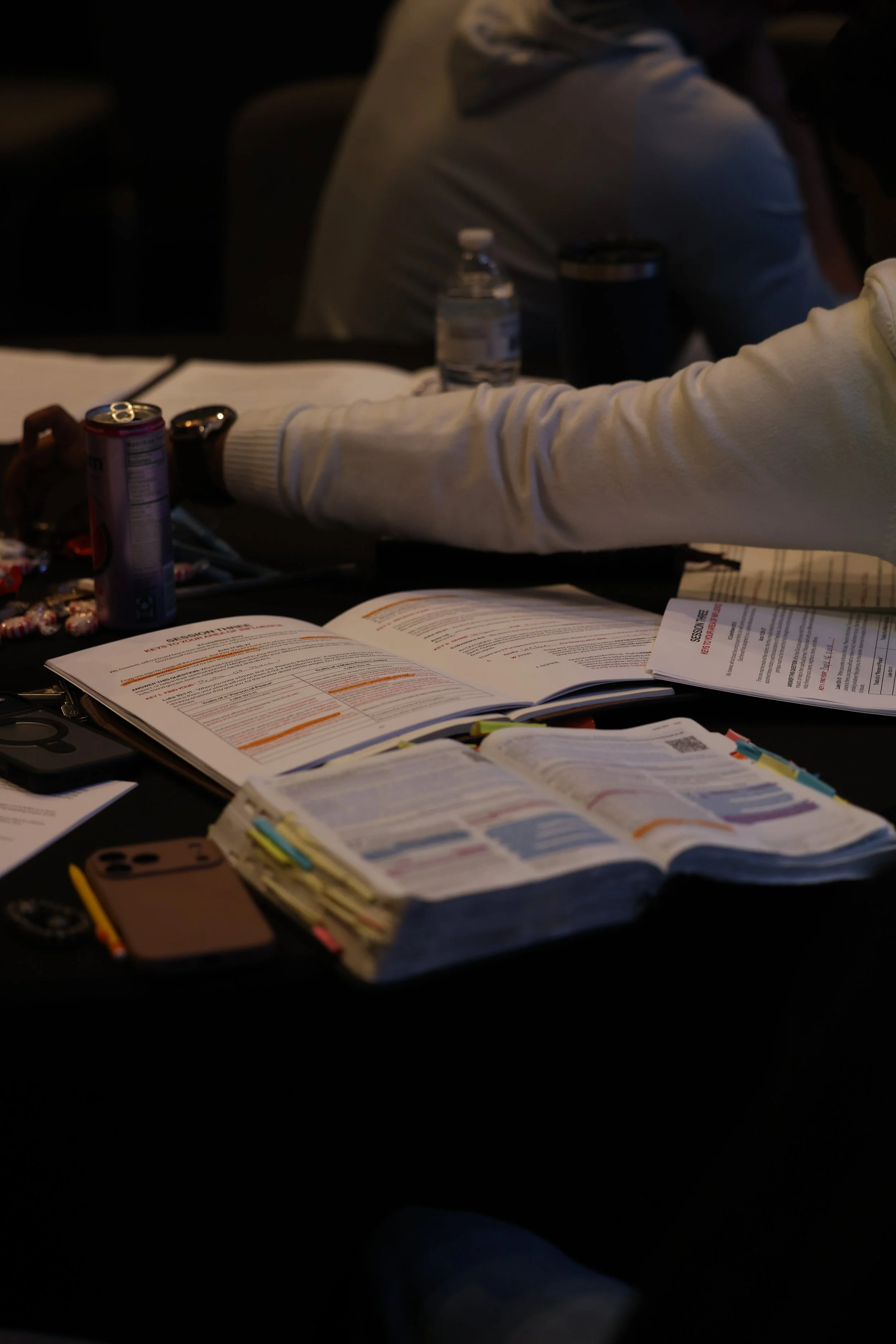 A person wearing a white long-sleeve shirt sitting at a table with open books, papers, a can, a water bottle, and a phone, in a dimly lit environment, possibly during a meeting or study session.