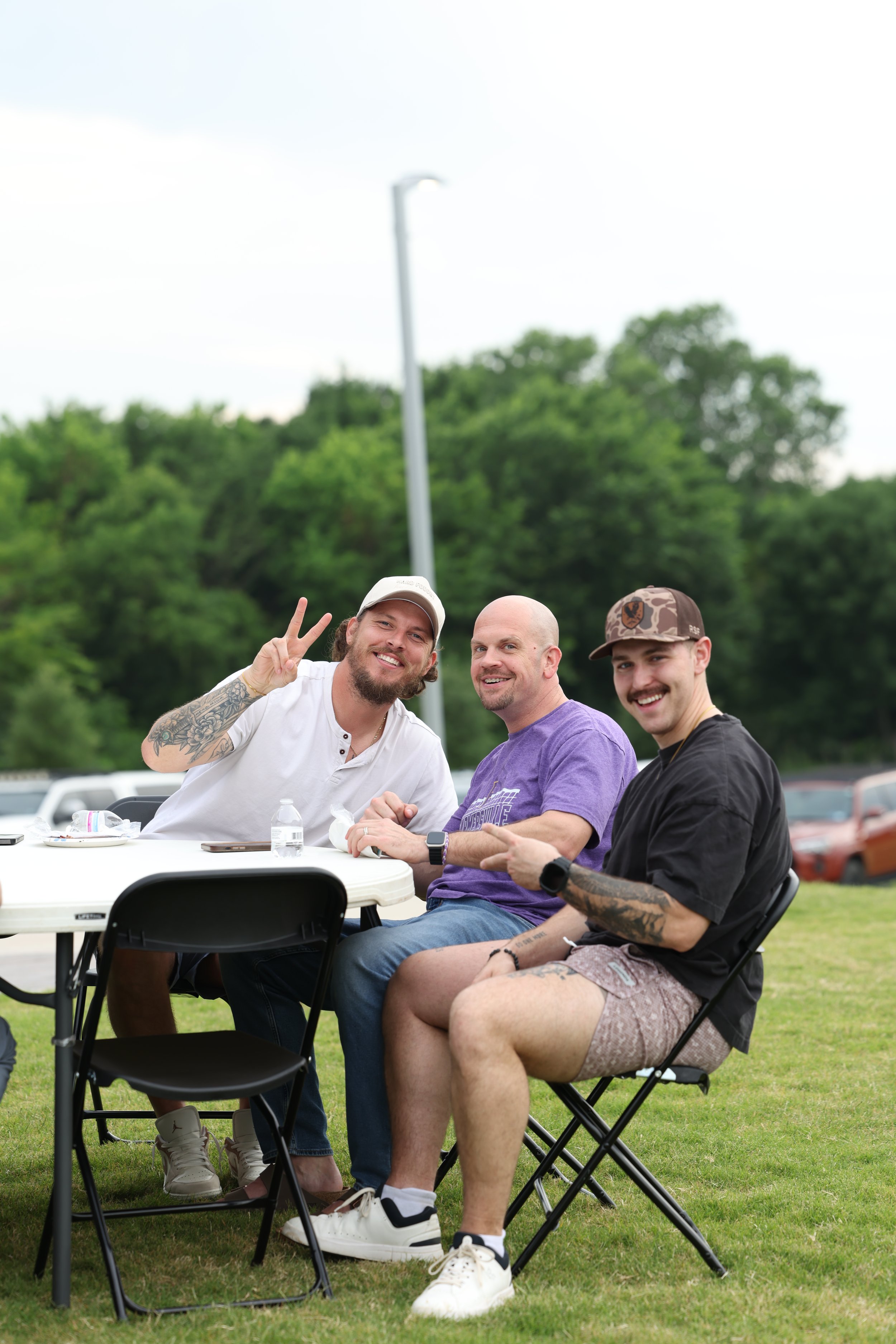 Three men sitting and smiling outdoors at a table on grass, with a background of trees and a cloudy sky. One man is making a peace sign.
