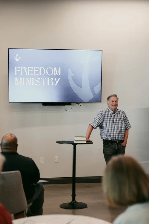 A man standing at a podium next to a table with books in front of a large screen that displays 'Freedom Ministry' and a logo in a conference room with seated audience members.