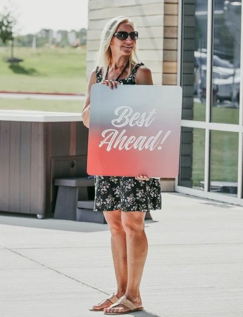Smiling woman holding a sign that says 'Best Ahead!' outside a modern building.